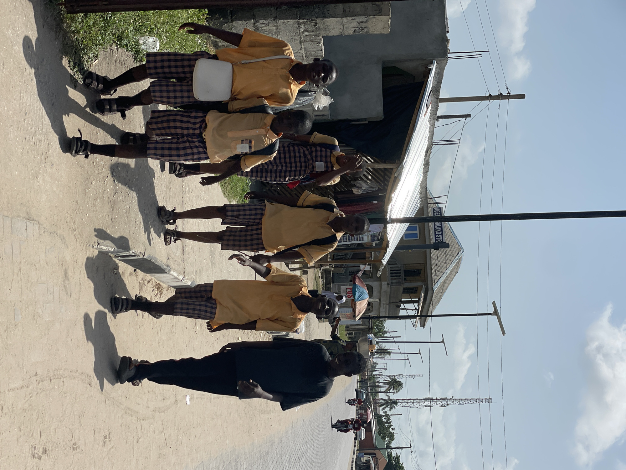 The image depicts a street scene in Badagry, Nigeria. A group of five people, likely students, are walking along a dirt road. The students are wearing school uniforms: yellow shirts and plaid shorts. One man is wearing a black t-shirt and black pants. He appears to be walking alongside the students, facing toward the camera. Behind them are buildings of various heights. Power lines and poles stretch across the sky, with a few palm trees visible in the background. The street is lined with low-lying structures, including some under construction.