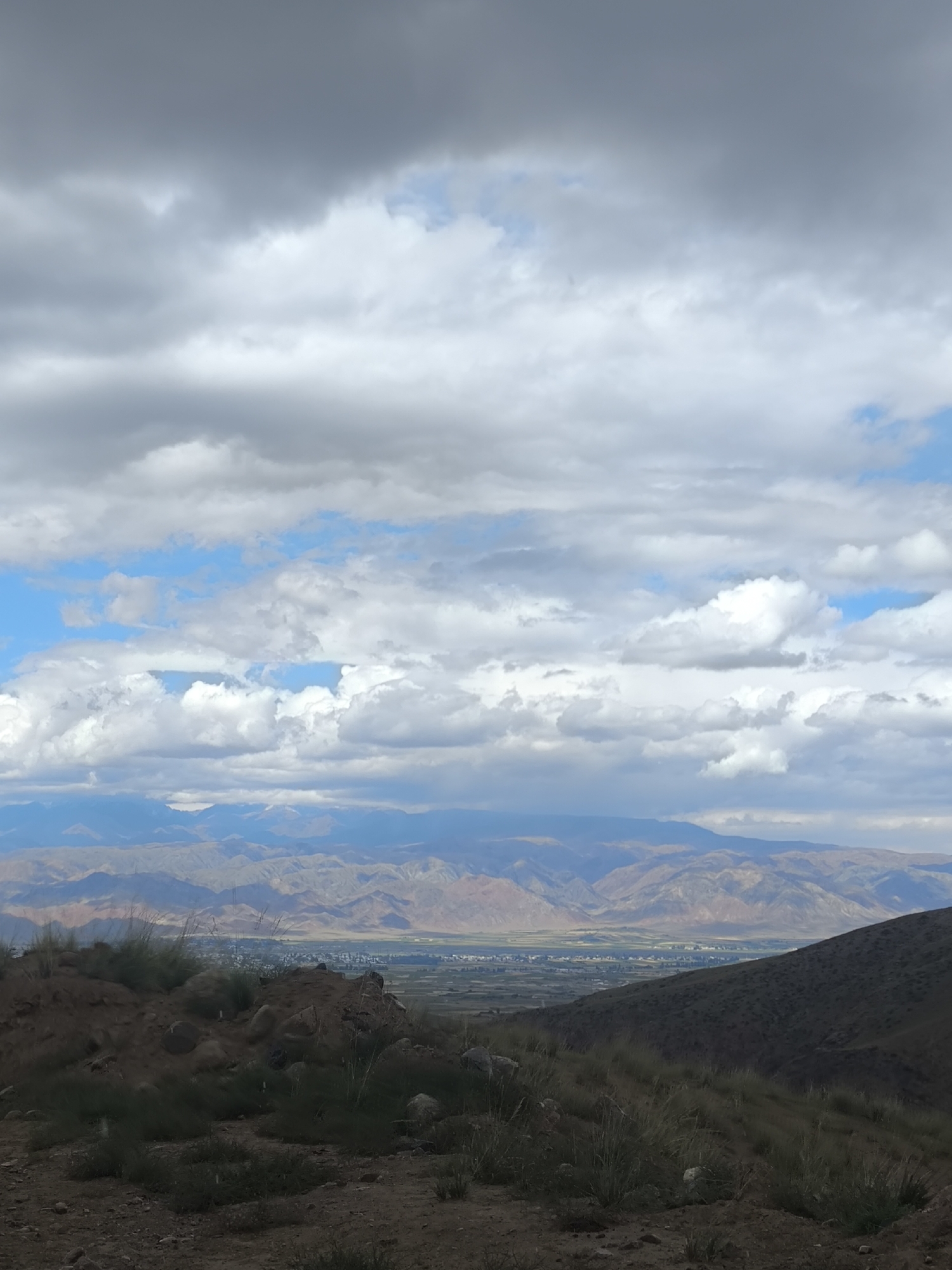 A high-angle, wide shot captures a vast mountain landscape under a cloudy sky, with a hint of a city visible in the distance. The foreground is dominated by dry, rocky soil dotted with sparse, dry grass and a few scattered rocks. A small, dark hill rises on the left, covered with similar vegetation. The middle ground reveals a valley with hints of human settlement, appearing as a cluster of structures and fields. This valley is bordered by rolling hills that transition into towering, layered mountains in the background. The mountains exhibit a muted palette of browns, grays, and purples, with some distant peaks showing hints of snow. The sky is filled with a dynamic mix of dark, heavy clouds and patches of bright blue, suggesting an overcast or partly cloudy day. The scene is devoid of any people or specific actions. The location is identified as being in the vicinity of the city of Bolshevik, Kyrgyzstan.