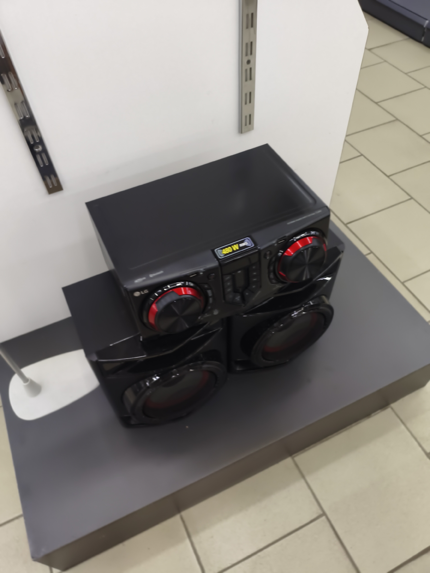An LG stereo system with two speaker units is displayed on a gray platform in front of a white wall with metal shelving. One speaker unit is stacked on top of another, with the main stereo unit positioned on the upper speaker. The stereo unit has red illuminated knobs and a display showing "480 W RMS". The scene appears to be indoors, possibly in a retail store or showroom, with light-colored tiled flooring visible in the background. No people are present in the image. The location is identified as Aktobe, Kazakhstan.