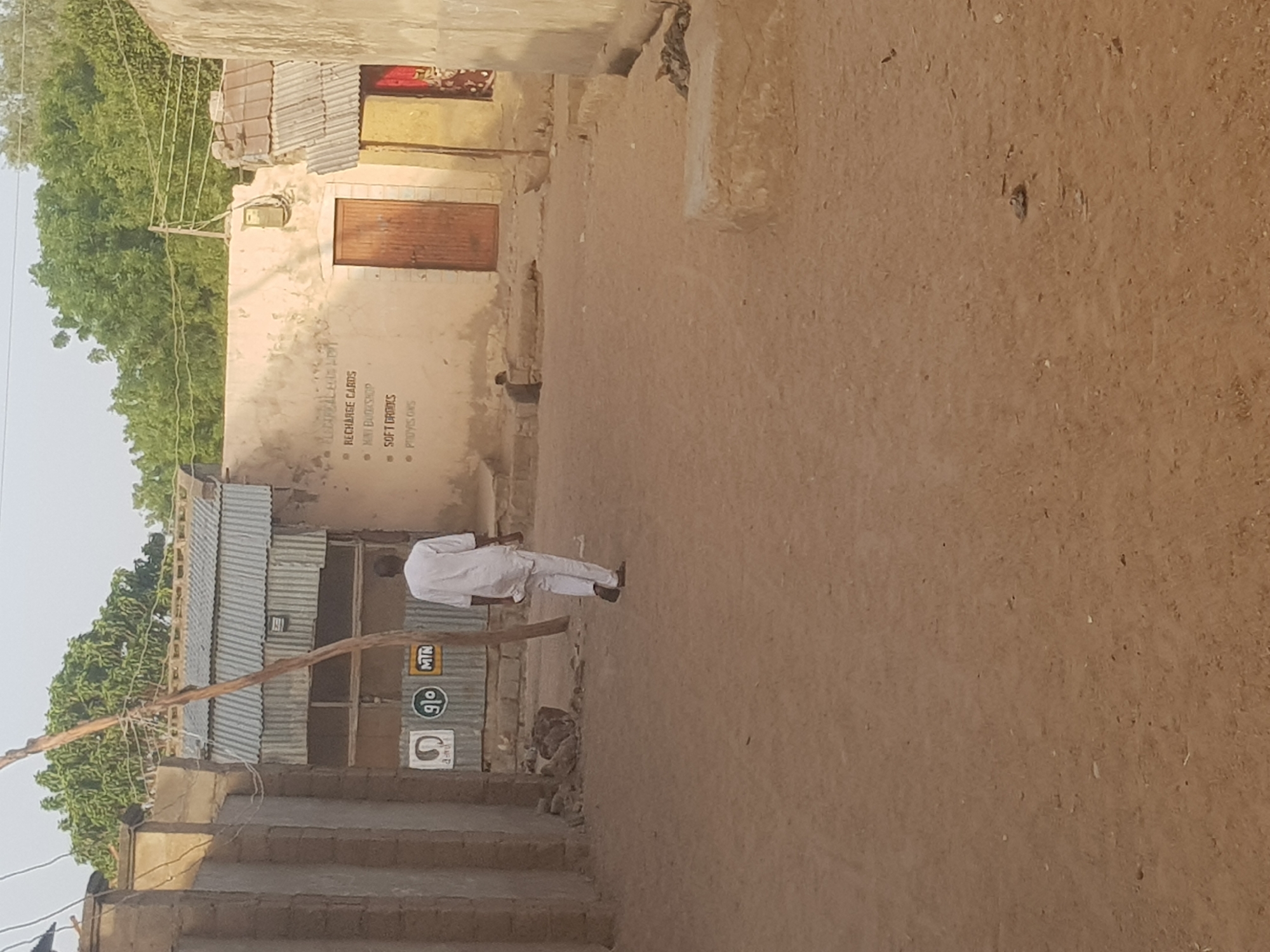 The scene is a dusty street in Dutsinma B, Nigeria. The environment appears arid with vegetation to the left. Buildings on both sides of the street have walls of earthen material with corrugated metal roofs. A man is walking along the street toward the right, wearing light-colored clothing. On the left, a building has signs for "Recharge Cards", "Soft Drinks", and "Provisions" painted on its exterior. Telecommunication signs are present on a building adjacent to it. The ground is a compacted dirt surface.