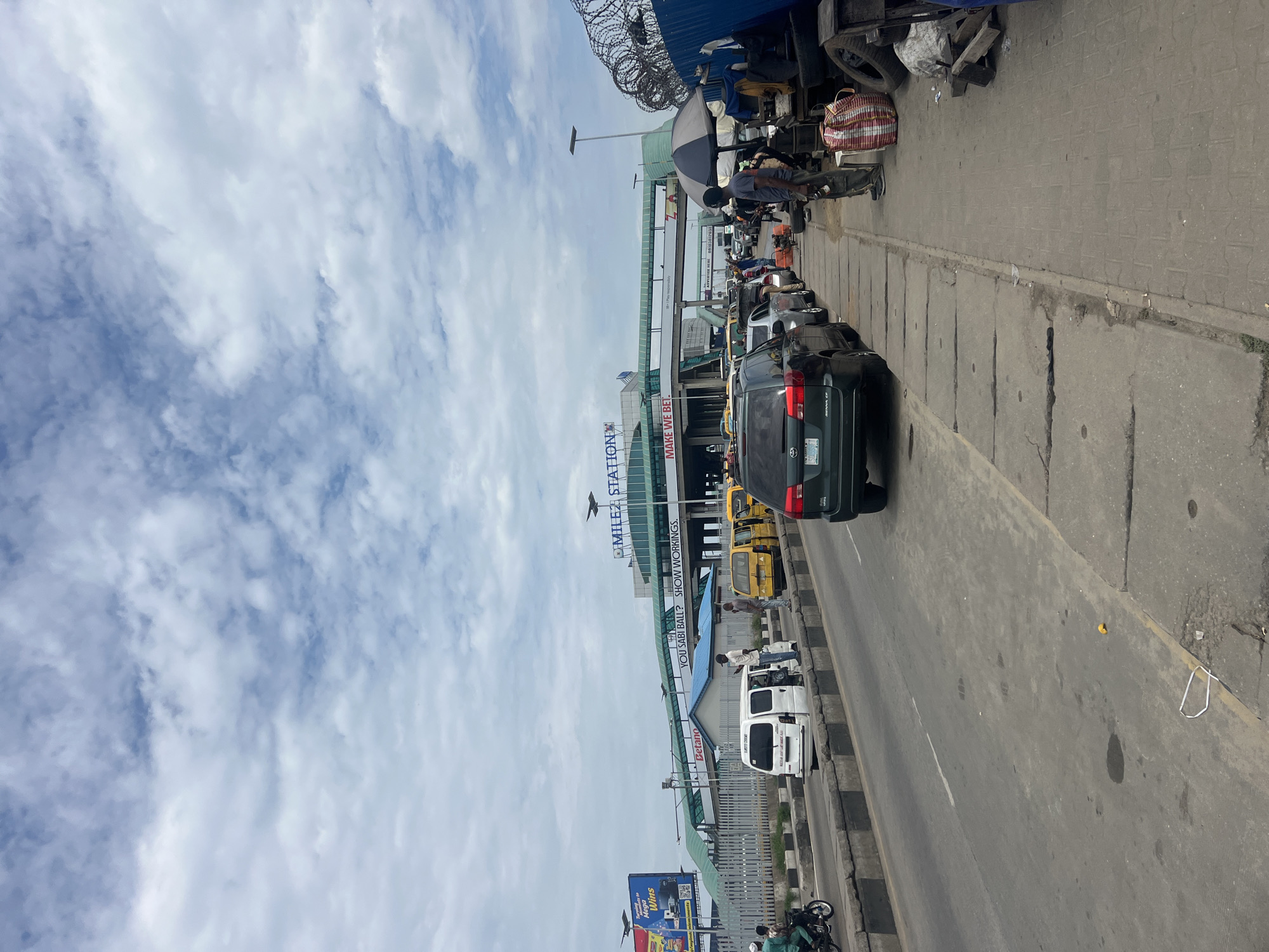 The image depicts a street scene in Agbani, Nigeria, featuring a busy road with various vehicles, including cars, vans, and yellow minibusses. A pedestrian walkway runs alongside the road, lined with stalls and informal vendors. In the background, a large building with "MILE 2 STATION" prominently displayed suggests a transportation hub. The sky is partially cloudy, indicating daylight. People are seen standing and walking, some engaged in activities related to the stalls. The environment appears to be urban, with paved roads and concrete sidewalks, and evidence of commercial activity.