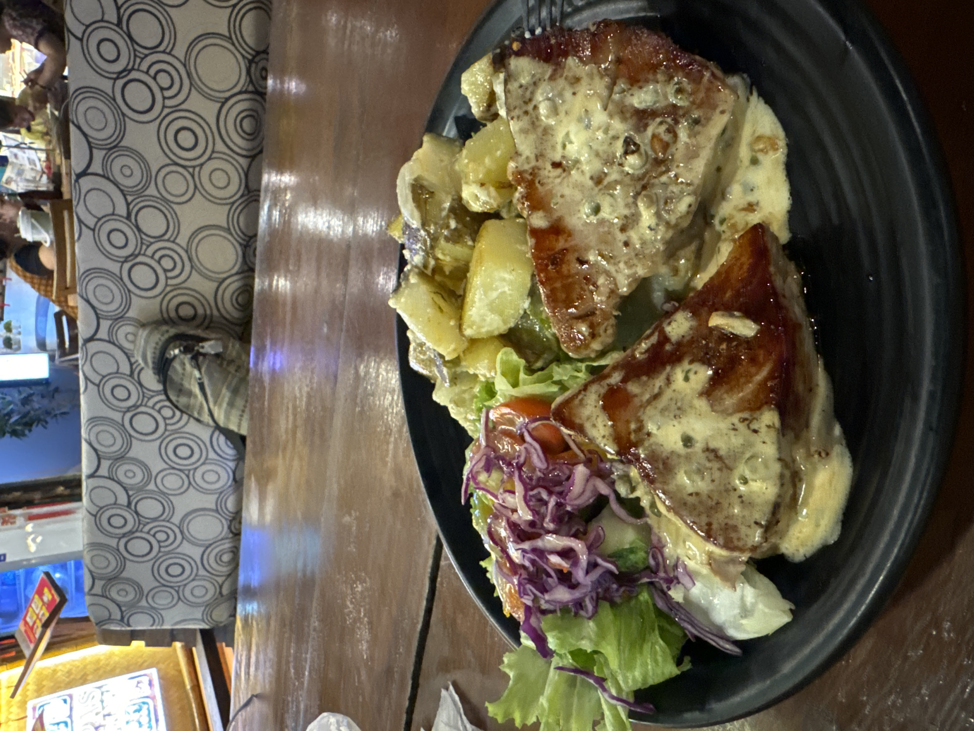 The image depicts a close-up, eye-level shot of a black plate of food on a wooden table. The plate contains two triangular pieces of pan-seared fish covered in a creamy sauce with visible herbs, accompanied by a side of potatoes with sauce and a salad with lettuce, red cabbage, and cherry tomatoes. In the blurred background, there are people seated on a patterned sofa, suggesting a restaurant or dining establishment. The setting appears to be indoors, with warm lighting. The overall impression is of a casual dining scene, likely in Mũi Né, Vietnam, given the context provided.