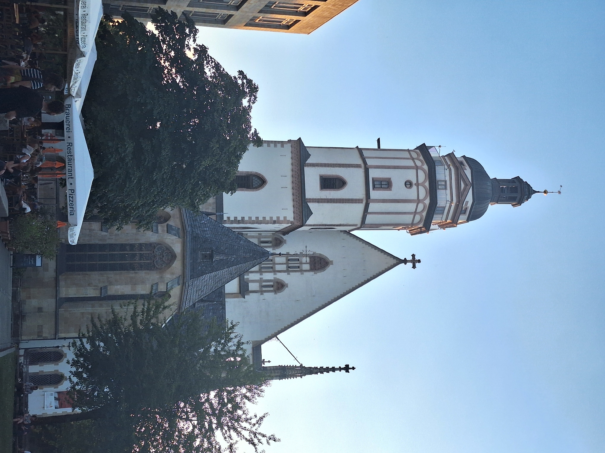 The image depicts the historic Nikolaikirche (St. Nicholas Church) in Leipzig, Germany, bathed in the soft light of what appears to be late afternoon or early evening. The towering Gothic church, with its distinctive white facade and ornate steeple, dominates the center of the frame. A large, dark green tree partially obscures the lower portion of the church. In the foreground, a row of white outdoor umbrellas, bearing the text "Brauerei • Restaurant • Pizzeria," shades a busy outdoor seating area. Several people are visible sitting at tables and walking around, engaged in dining or socializing. The sky is a clear, pale blue, indicating fair weather. To the left of the church, a modern building with large windows is visible, contrasting with the historical architecture.