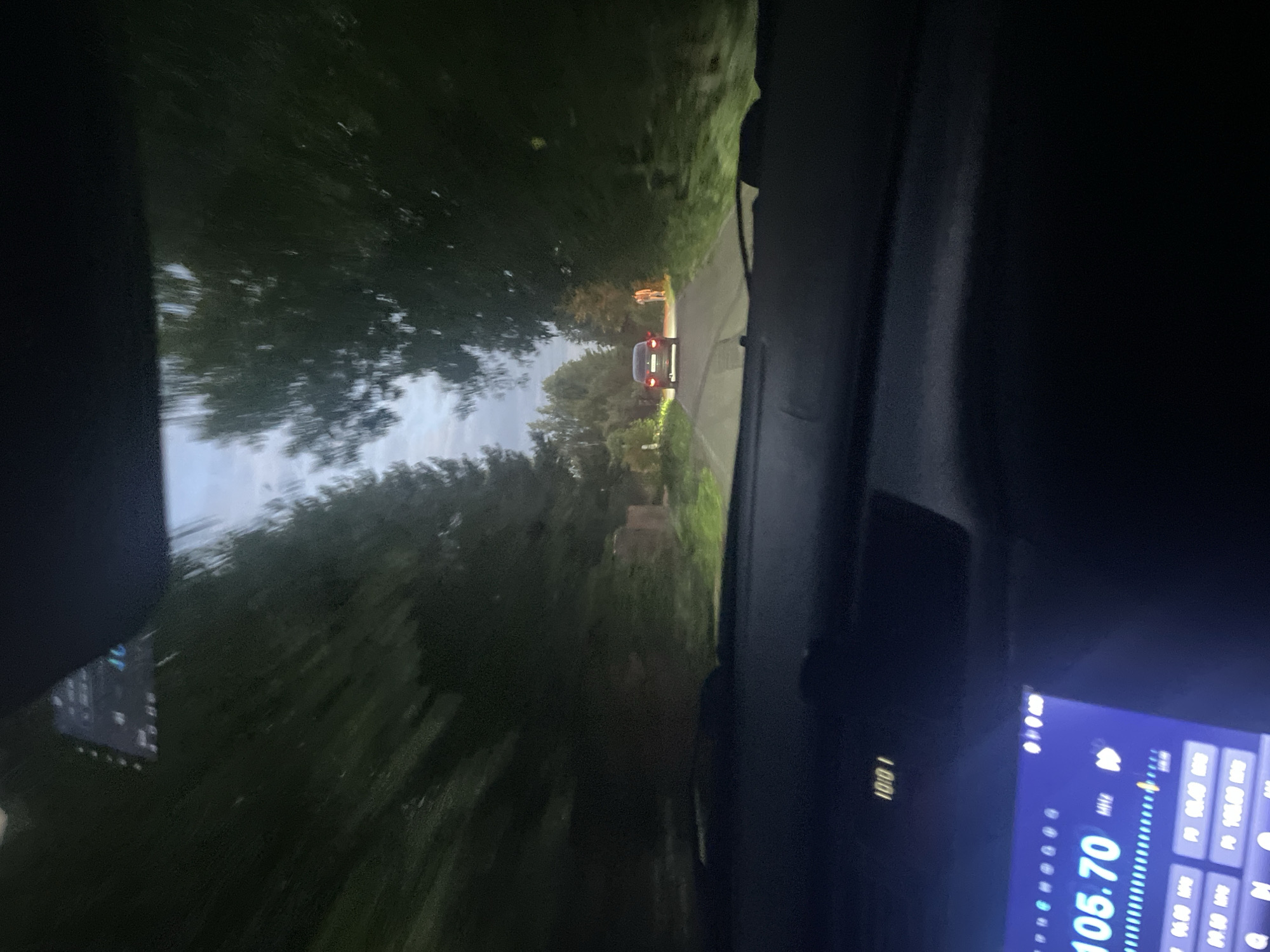 The image captures a nighttime view from inside a vehicle driving on a paved road lined with trees. The road appears to be in a semi-rural or suburban area, possibly on the outskirts of Vnukovo, Russia, given the context provided. In the distance, a dark-colored car is visible, moving away from the viewer. To the right of the road, illuminated by the car's headlights or ambient light, are two figures. One figure, standing by the roadside, appears to be wearing light-colored clothing. The other figure is less distinct. The dashboard of the vehicle is partially visible, displaying a digital clock that reads "10:01" and a radio interface with "105.70 MHz" prominently displayed. The overall lighting suggests it is dusk or nighttime, with artificial lights from the car and possibly streetlights or house lights contributing to the illumination of the scene. The trees on either side of the road create a somewhat enclosed tunnel effect.