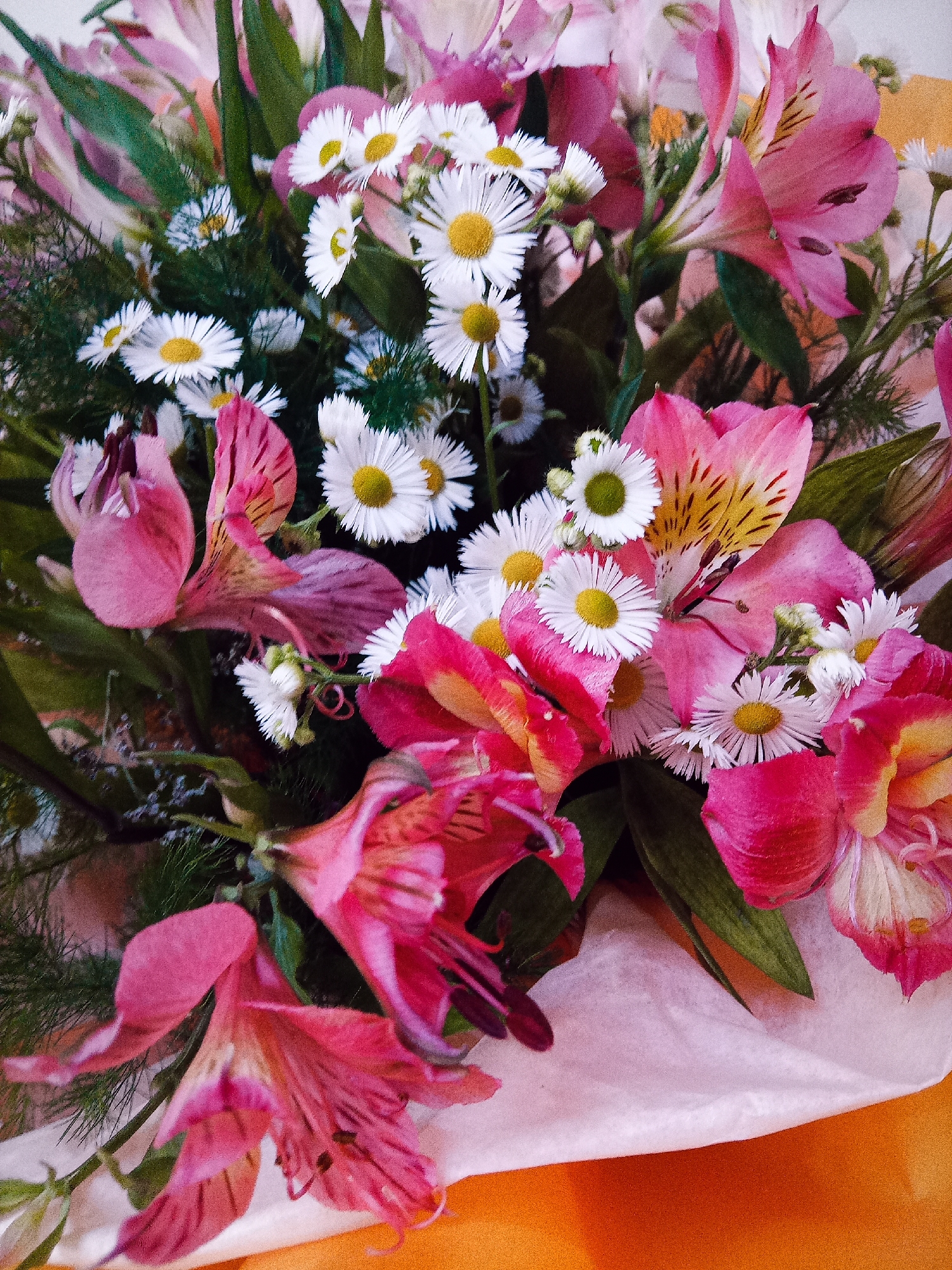 The image shows a close-up of a bouquet of flowers. The bouquet contains a variety of flowers, including pink alstroemeria and small white daisies with yellow centers. Some of the alstroemeria flowers are fully bloomed, while others are still in bud. The flowers are arranged in a lush, dense bouquet, with green foliage visible throughout. The bouquet is wrapped in white tissue paper, and a portion of an orange surface is visible beneath it. The image appears to be a still life arrangement, with no people or discernible actions taking place. There is no information in the image to suggest a specific location in Chișinău, Moldova.