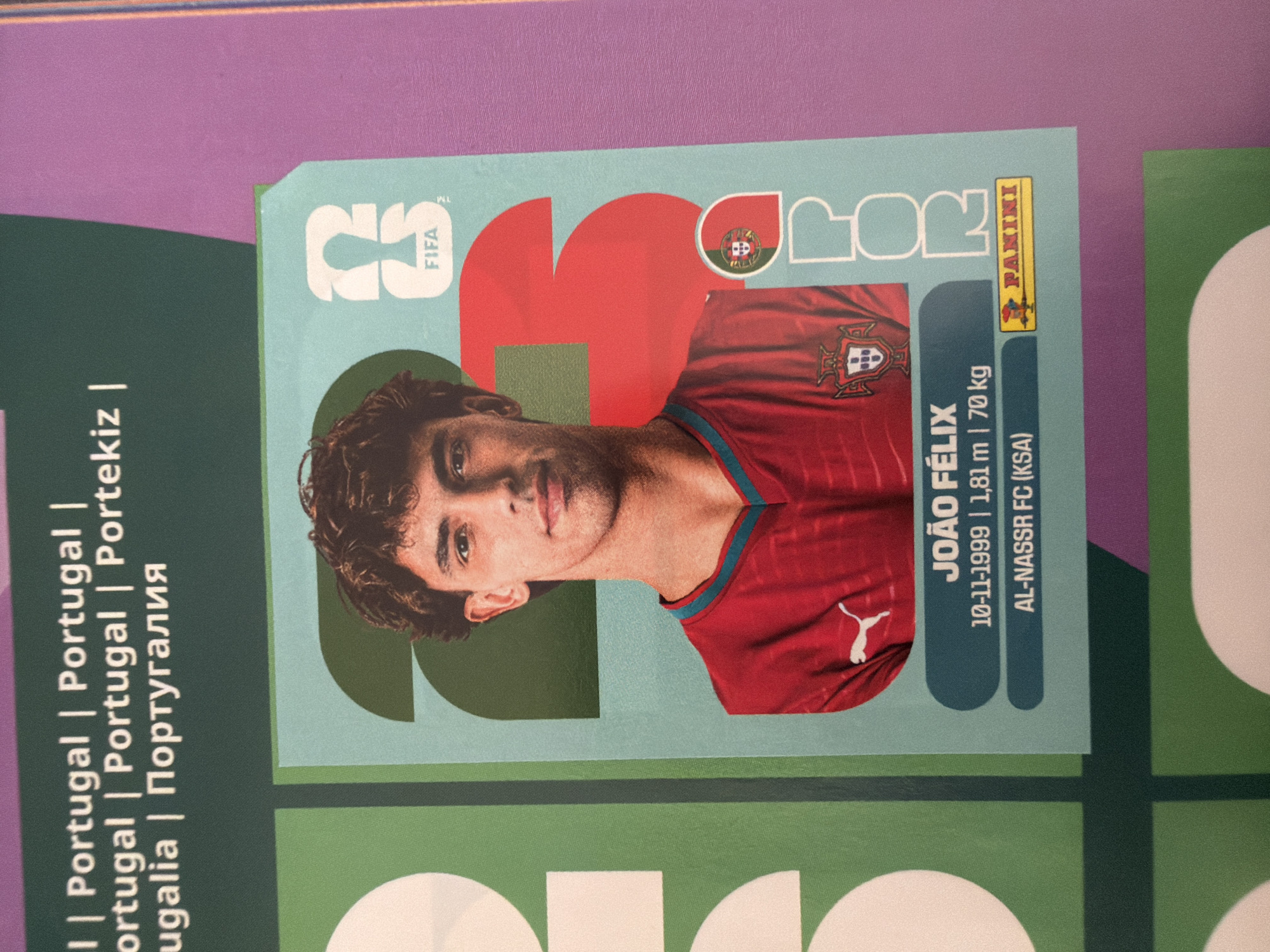 The image is a close-up of a trading card featuring Portuguese footballer João Félix. The card has a light blue background with large red numbers "23" on it. João Félix is wearing a red jersey with the Portuguese crest and a white Puma logo. Below his image, his name "JOÃO FÉLIX" is printed, followed by his birthdate "10-11-1999", height "1,81 m", weight "70 kg", and current club "AL-NASSR FC (KSA)". The Panini logo is visible at the bottom right. The card is part of a larger collection, with parts of other cards and a purple background visible. The image does not provide any information about the city of Zlatitrap, Bulgaria, or any context related to that location.