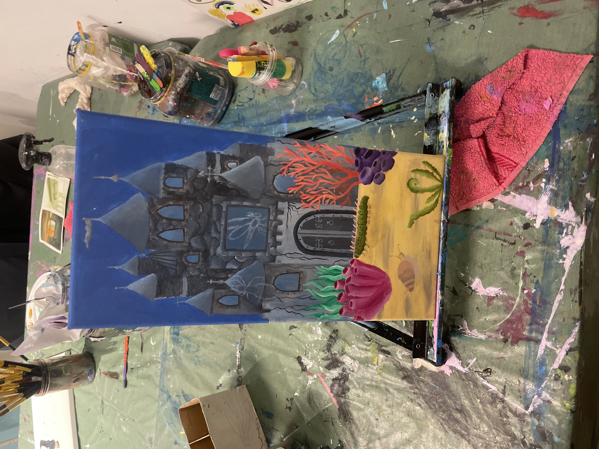 A close-up photograph shows a canvas painting in progress, likely situated in a studio or art space. The painting depicts a castle blending into an underwater scene, with the left side showing a grey castle against a blue background. The right side features a yellow background with painted coral, seaweed, a caterpillar, and a snail. The painting is placed on a black easel. The background is a cluttered green surface covered in paint splatters, with jars of paintbrushes and other art supplies visible. A pink towel sits on the right side of the easel. The location is Raanana, Israel.