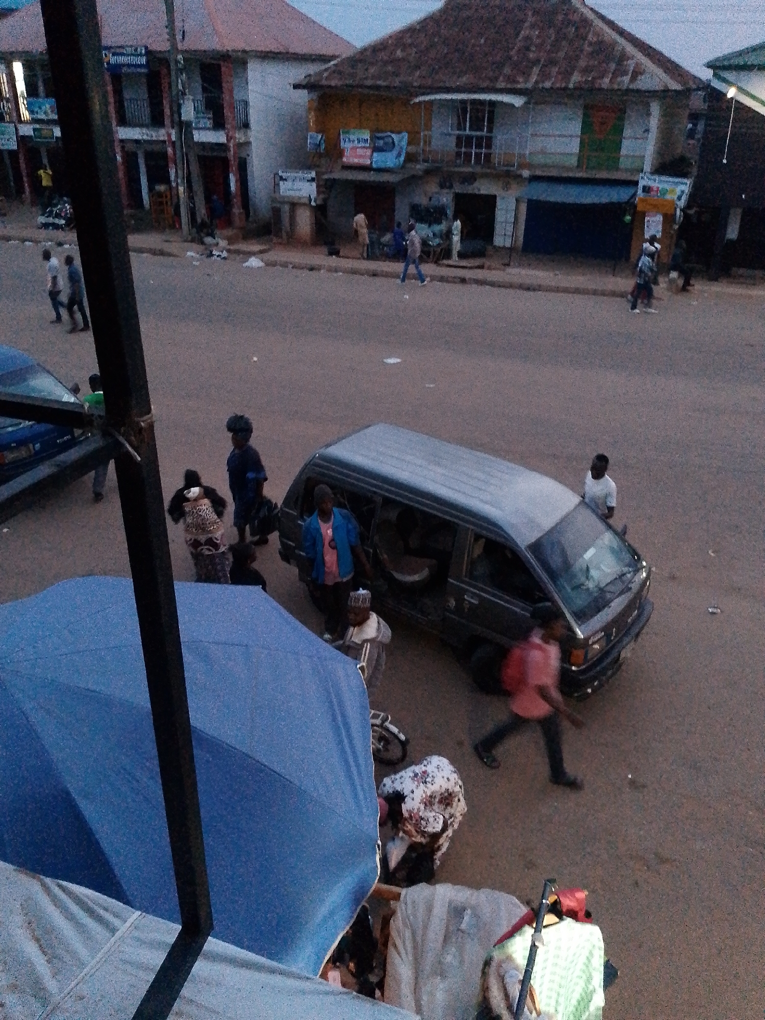 The image depicts a street scene in Bukuru, Nigeria, with a mix of people, vehicles, and buildings. In the foreground, a large blue umbrella dominates the lower left portion of the frame, with a light gray tarp visible beneath it. A dark, vertical pole extends upwards from the umbrella.
In the mid-ground, several people are visible. A dark-colored van is parked on the right side, with its passenger door open. A man in a blue jacket and pink shirt stands near the open door. Another man, wearing a light-colored shirt and dark pants, is seated on the ground near the van. Further to the left, a woman in a patterned dress and headwrap is partially visible, appearing to be interacting with someone or something out of frame. A woman in a dark blue dress and headwrap stands with a bag in her hand. A man in a white shirt and dark pants is walking away from the van.
In the background, a building with a reddish-brown roof is prominent, featuring multiple windows and balconies. Several shops with shuttered entrances are visible along the street. Other individuals are scattered throughout the scene, some walking, others standing. A blue car is parked on the far left. The street itself appears to be paved, with scattered debris on the ground. The overall lighting suggests it is either early morning or late afternoon.