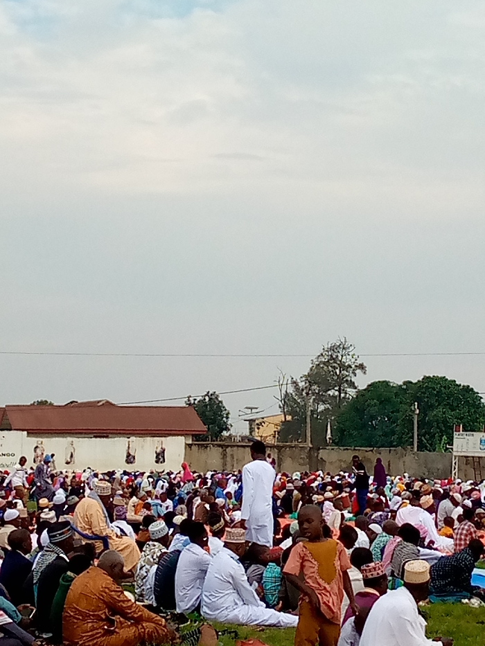The image shows a large gathering of people in an outdoor area in Beni, Democratic Republic of Congo (coordinates: 0.489, 29.463). The people are sitting on the ground, mostly facing towards the right side of the image, likely participating in a religious gathering. Many of the men are wearing white clothing, and some are wearing hats. There is a low concrete wall in the background with a brick building behind it. Trees and a cloudy sky can be seen in the background. There is an information board visible to the right with a sign that reads "PLAISTY MAISTER". The people are facing towards the right, but the viewer's perspective is facing them from behind. The grassy area is sparsely covered with vegetation.
