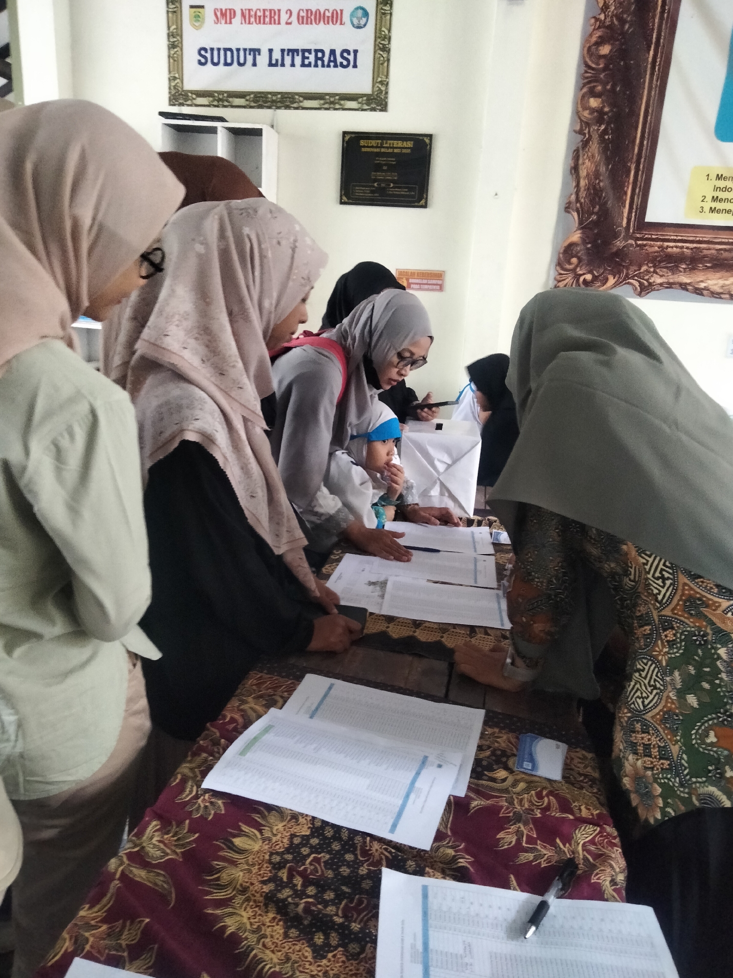 In a classroom or public building with a sign that reads "SMP NEGERI 2 GROGOL SUDUT LITERASI," several people are gathered around a table covered with a patterned cloth. The people, who appear to be students or participants, are mostly wearing headscarves (hijabs). They are looking down at papers and some are interacting with each other. One person is holding a pen, suggesting they are writing or filling out forms. There are stacks of papers and a small white card on the table. The environment seems to be indoors, with a wall in the background and decorative framing on the wall. The overall scene depicts an organized activity, possibly related to literature or a school event. The specific location within Sanggrahan, Indonesia, is not directly discernible from the image itself, but the sign indicates it is a school.