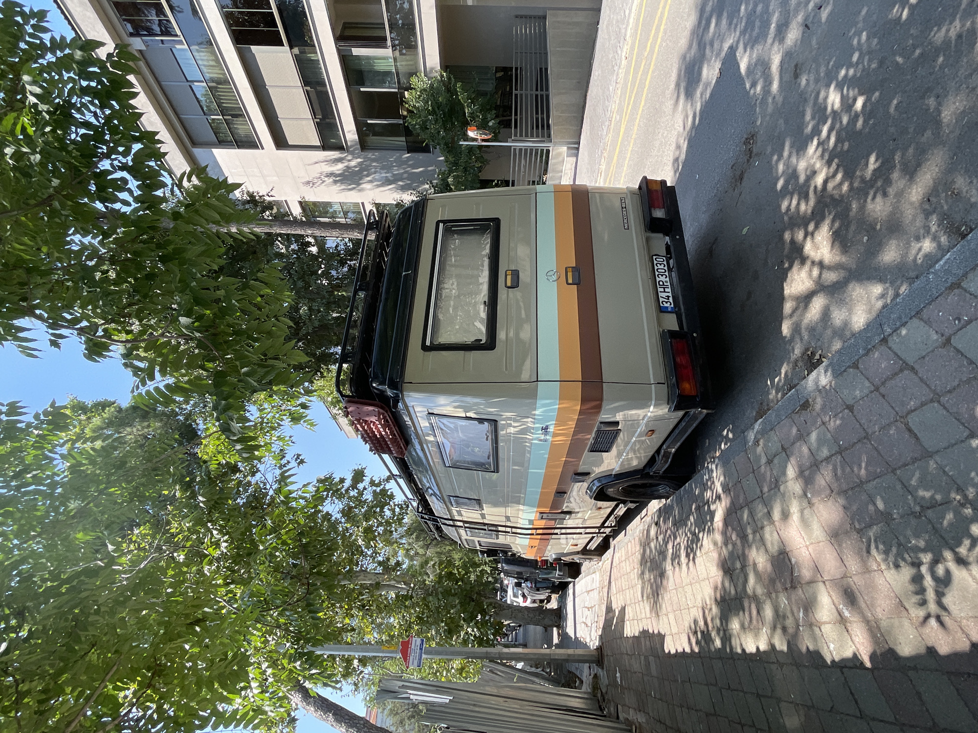 A beige Mercedes-Benz camper van with orange and light blue stripes is parked on a paved street in Kadıköy, Turkey. The van has a roof rack with a red traction mat. The street is lined with trees casting shadows on the sidewalk and road. In the background, a modern building with balconies and large windows is visible. The license plate of the van reads "34 HP 3030". The scene appears to be on a sunny day.