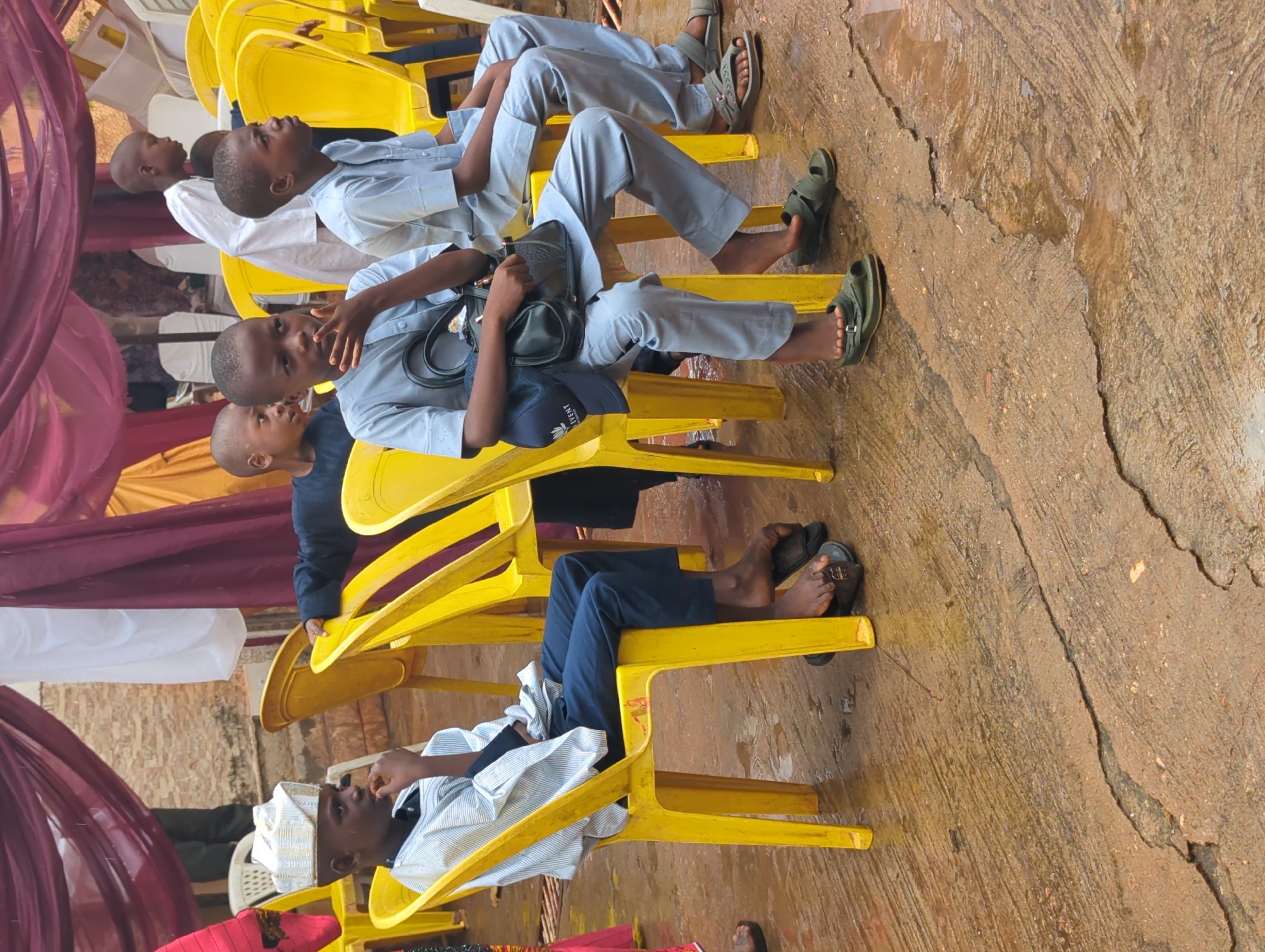 The image captures a group of young males, likely boys or teenagers of African descent, seated on yellow plastic chairs in what appears to be an outdoor or semi-outdoor setting in Unguwar Rimi, Nigeria. The image is oriented vertically.
The subjects are dressed in light-colored, long-sleeved shirts and trousers, mostly in shades of light blue or grey, with one boy wearing dark blue trousers. Some are barefoot or wearing sandals/slides; one black sandal prominently displays a white "B" logo. One boy wears a white embroidered cap (kufi), and another has a black shoulder bag resting on his lap, over which a dark blue cap with partially visible white text, possibly "STRIENT" or similar, is placed.
The scene suggests a gathering or event, indicated by the arrangement of chairs and decorative fabric drapes in maroon/purple, yellow, and white visible on the left side of the frame. The boys are mostly looking forward or slightly upwards, appearing attentive or observing something out of frame. The ground beneath them is a worn, textured concrete or paved surface, showing cracks and what might be dampness or stains.
The bright, natural lighting indicates it is daytime. The overall atmosphere is calm, with no strong emotions discernible.