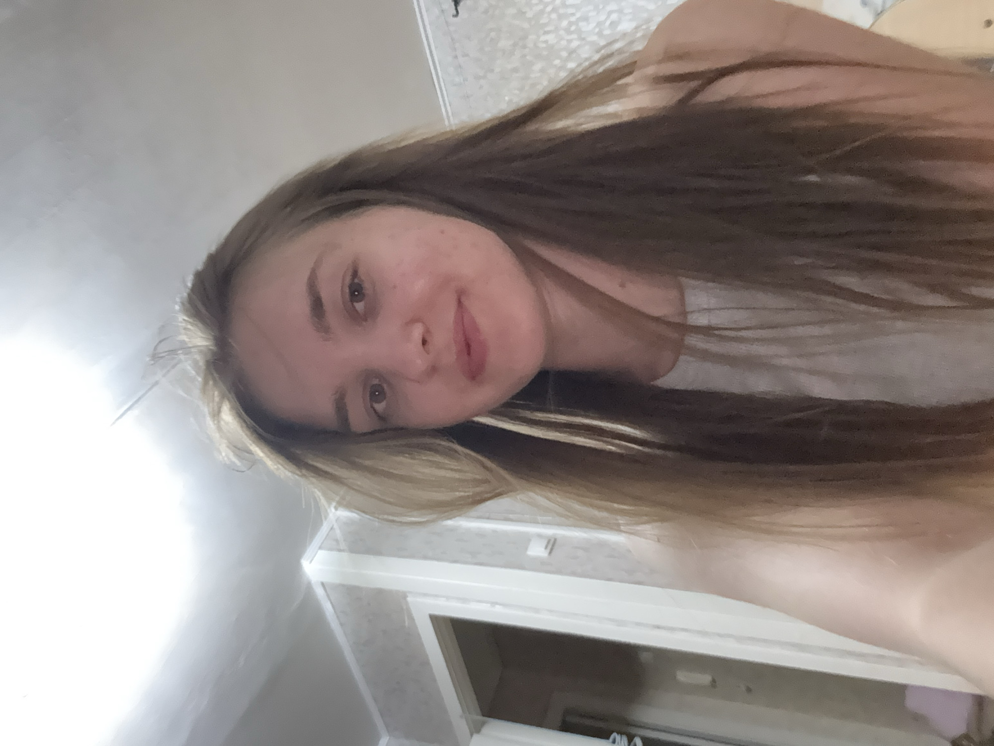A woman with long brown hair is facing the camera, smiling slightly. She appears to be indoors, judging by the wall and doorframe visible in the background. The wall is a light color and the doorframe is white. The location is unclear, and there is no evidence to suggest it is in Perm, Russia.