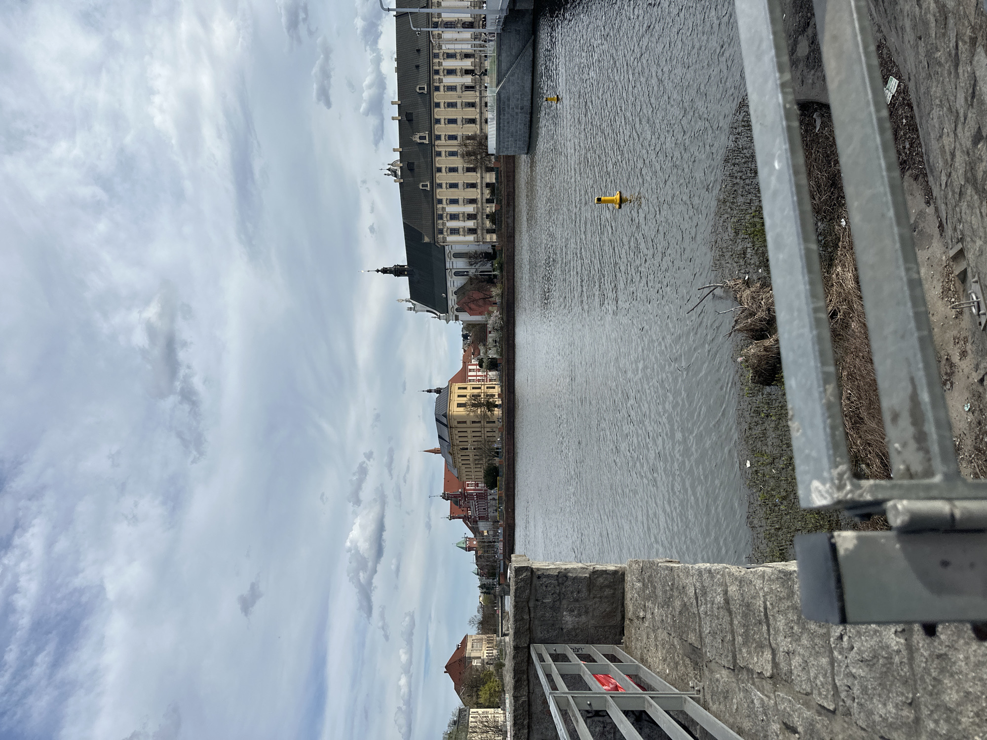 The image captures a view of Wrocław, Poland, from a bridge overlooking the Oder River. The sky is partially cloudy. Buildings line the riverbank, including the University of Wrocław with its distinctive baroque architecture. Two yellow buoys are visible in the water. A metal railing and a stone wall of the bridge are in the foreground, partially obscuring the view. The environment appears calm and urban. No people are visible.