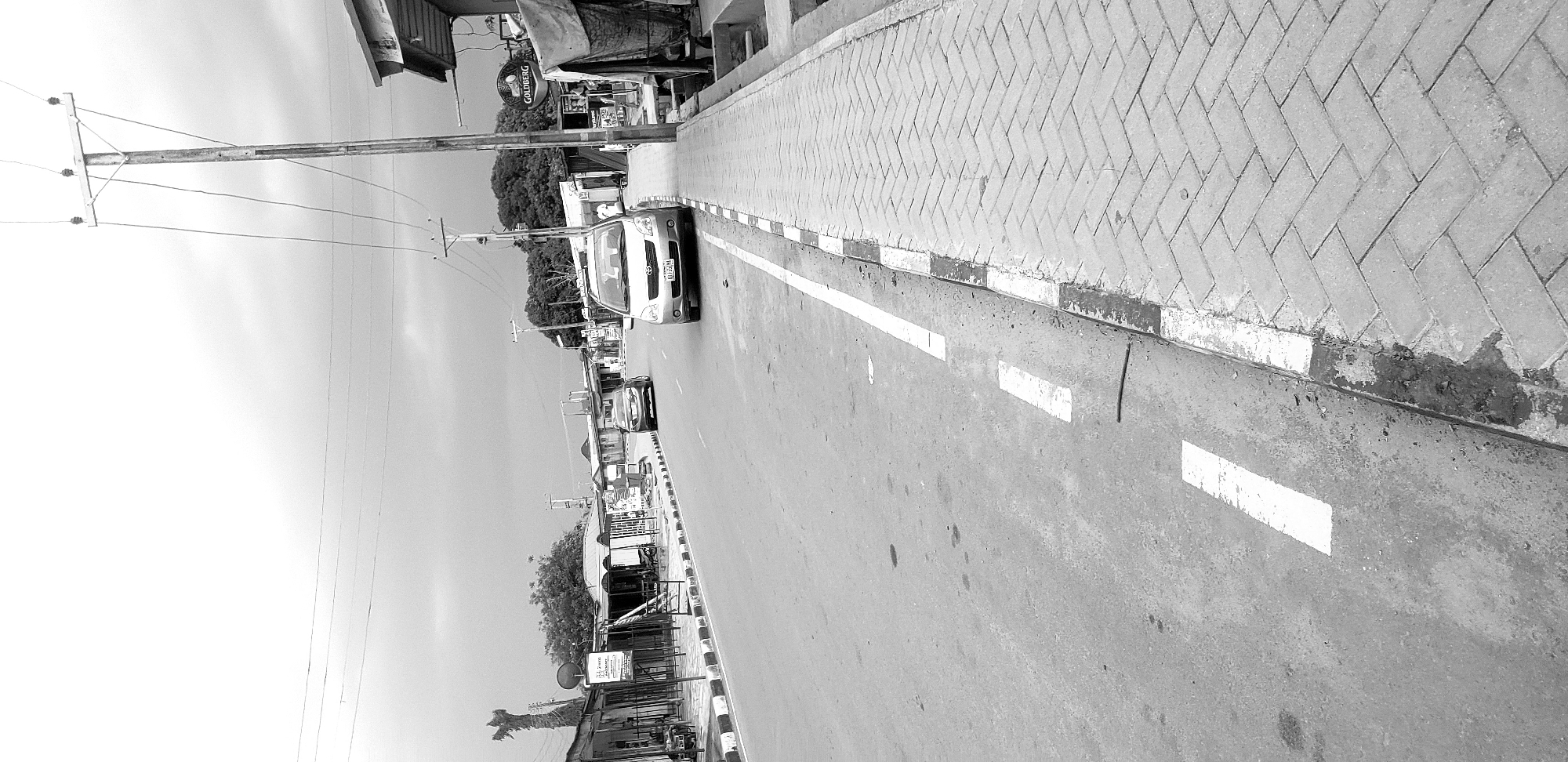 This black and white photograph depicts a bustling street scene in Narayi, Nigeria. Although the original image is oriented vertically, when viewed correctly, the paved road stretches into the distance, flanked by a sidewalk and various buildings.
The asphalt road features clear white dashed lane markings. To the right, a wide pedestrian sidewalk is constructed from light-colored paving bricks laid in a distinctive herringbone pattern. A curb, painted with alternating black and white stripes, separates the road from the sidewalk.
Vehicles are present on the road, with a prominent light-colored sedan (likely a Toyota, bearing the partial license plate "KD10-910") in the mid-ground, heading away from the viewer. Another darker vehicle can be seen further down the street. Utility poles, laden with multiple overhead wires, line the left side of the road, receding into the background.
The street is lined with various structures, appearing to be shops or commercial establishments, including one with a visible round "GOLDBERG" sign. Trees are interspersed among the buildings. The sky is bright and appears partly cloudy, indicating daytime. The scene captures an ordinary moment of daily life in a town or city, with general traffic movement and commercial activity suggested by the shops. A black cable or hose is visible on the curb in the foreground, and some marks are present on the road surface.