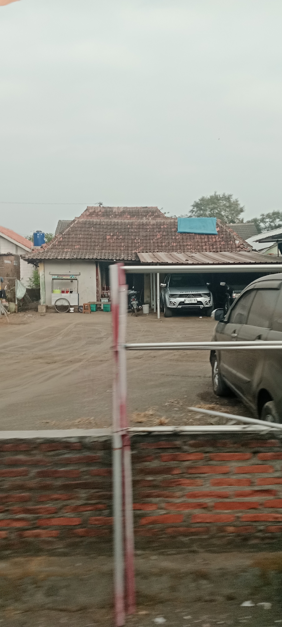 The image shows a street scene in Maguwoharjo, Indonesia, with a building that appears to be a small business or residence. The building has a tiled roof and a covered parking area where several vehicles are parked, including a silver SUV with a license plate that reads "N 1297 EI" and a darker colored SUV. There is also a motorcycle parked under the cover. In front of the building, there is a food cart with various containers of food and drinks. The ground is dirt and appears to be a roadway or open area. In the foreground, there is a red and white striped pole and a brick wall, suggesting the image was taken from a vehicle or a roadside barrier. The sky is overcast, and the overall lighting suggests it is daytime, possibly early morning or late afternoon.