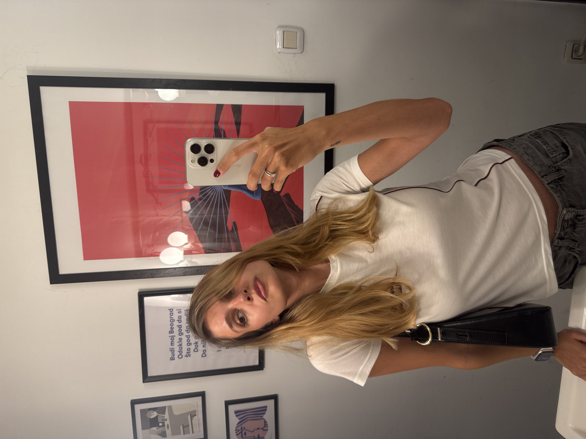 A woman with blonde hair takes a selfie in a bathroom. She is wearing a white t-shirt and gray denim shorts. She is holding a white smartphone in her right hand. A black purse is slung over her left shoulder. Behind her, on the wall, are several framed pictures. One large frame contains a red and black abstract print. Another frame, smaller, has text in a dark color on a white background. The visible text reads "Budi moj Beograd," "Odakle god da si," "Šta god da radiš," "Dokle god," and "Da ni." The setting appears to be a well-lit, modern bathroom. The time of day and weather are not discernible. The woman's expression is neutral. The location context is Belgrade, Serbia.
