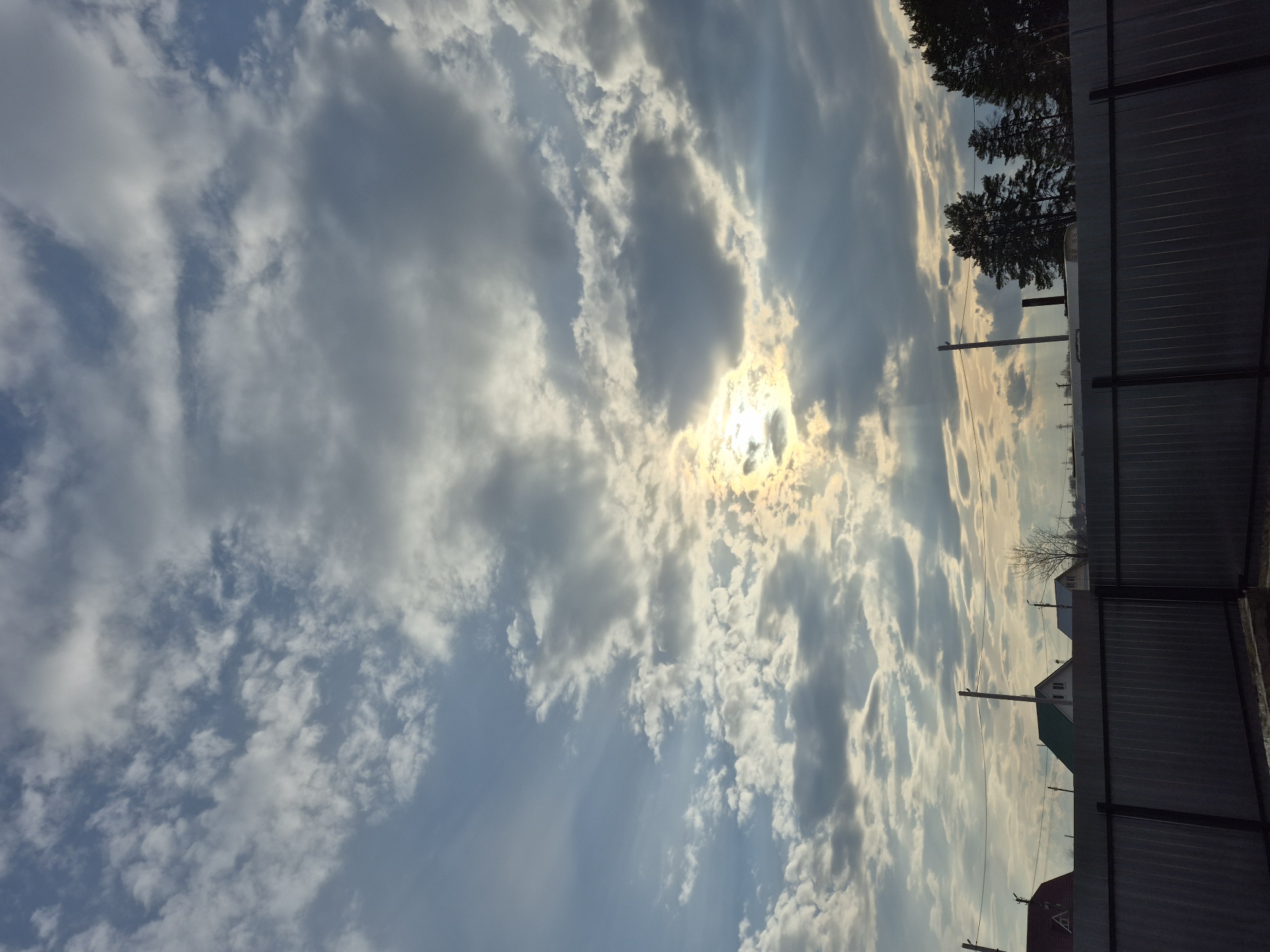 The image shows a sky filled with numerous clouds of varying size and density. Sunlight is visible through the clouds, creating a diffused, bright area in the sky. Part of a fence is visible on the right side of the image. Beyond the fence are trees, power lines, and the rooftops of several houses. The image does not contain specific information to determine the exact location in the city of Chaikovsky, Russia. No people are visible in the image. The environment is outdoors during daylight hours.