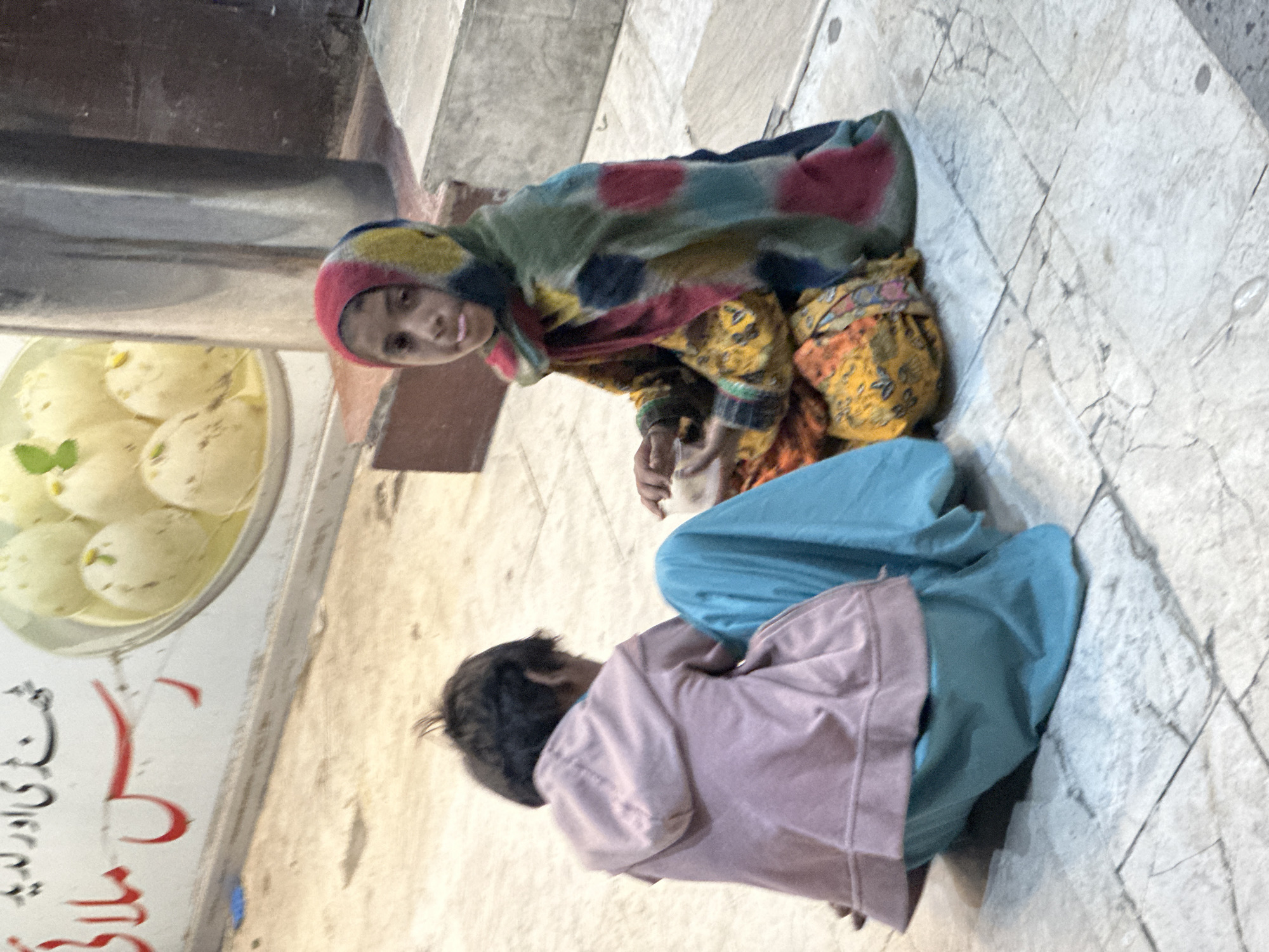 Two young individuals are seated on a tile surface. One individual, adorned in a colorful shawl and vibrant patterns, directs their gaze at the viewer. The other individual wears a light pink garment and light blue pants. Beside them is a display of desserts that appear to be Rasgulla. The sign next to the dessert case says "حجّی اور ریسی ملا". The location appears to be in front of a food shop.