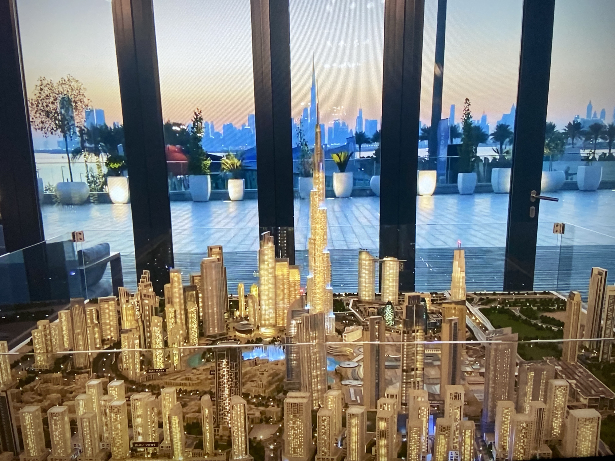The image shows a detailed architectural model of a city, dominated by numerous skyscrapers and urban structures. The model city features a central tall, slender tower resembling the Burj Khalifa, suggesting the location could be Dubai. The model includes roads, landscaping, and water features.
In the background, visible through a window, is an actual cityscape at sunset, with silhouettes of buildings and a similar prominent tall tower. The real cityscape is on a raised platform area featuring potted plants, suggesting a balcony or terrace view.
The model is placed in an indoor environment, possibly a sales office or exhibition space, as indicated by the architectural model and the upscale terrace view.