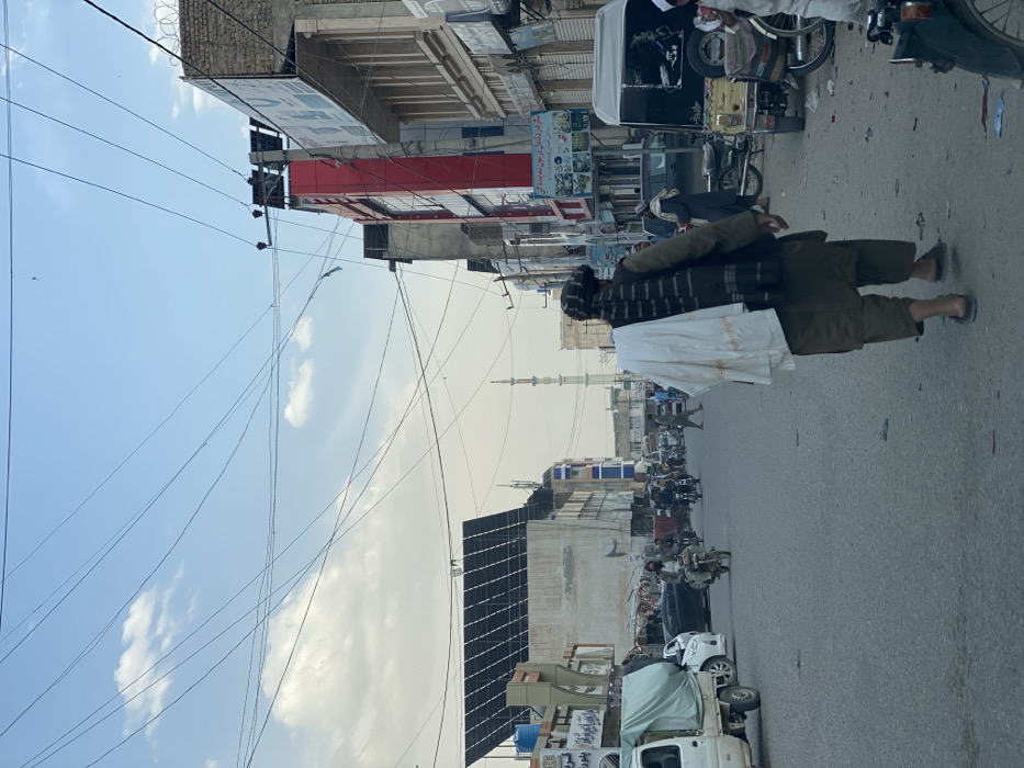The image depicts a daytime street scene in an urban area of Kandahar, Afghanistan, under a partly cloudy sky crisscrossed by numerous tangled overhead utility wires. The paved asphalt street shows signs of wear.
In the foreground, a male individual, viewed from the back, walks away from the camera towards the center of the street. He wears a dark head covering (turban), a dark vest over a lighter long-sleeved garment, and loose-fitting trousers (shalwar), with sandals on his feet. A white piece of fabric or garment is draped over his left shoulder.
To the left, a white light truck with a green tarp covering its bed is visible, followed by a white passenger car. Further down the street, multiple motorcycles, some with riders, and several pedestrians are present. On the left side of the street, multi-story buildings line the road. One building features a prominent, large array of dark-colored solar panels on its roof. In the distant background, a slender, light-colored minaret is visible.
On the right side of the street, a series of multi-story commercial buildings are present, one featuring a prominent red facade. Several buildings display advertisements and signs with text in Dari or Pashto. Barbed wire is visible on top of some of these buildings. A three-wheeled auto-rickshaw (tuk-tuk) is parked on the right curb, and a portion of another motorcycle is visible in the immediate far-right foreground.