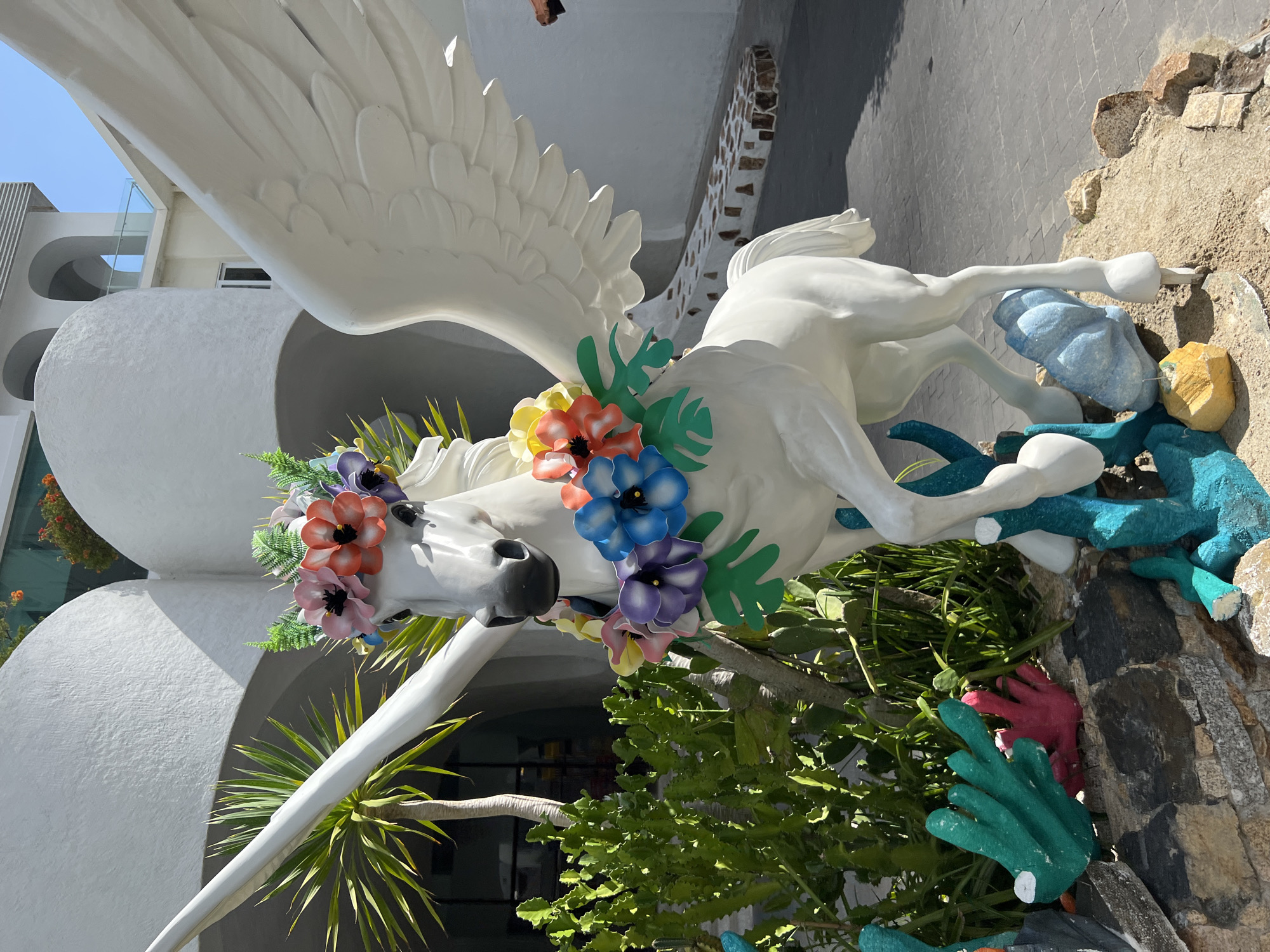 A white statue of Pegasus, a winged horse, is adorned with a vibrant floral wreath on its head and a garland of flowers around its neck. The statue is positioned outdoors in front of a white, modern building with curved architectural elements. To the left of the statue, a tall palm tree and a large, leafy cactus are visible. The ground is covered with sand and rocks, and there are also colorful, sculpted elements resembling coral in the foreground. The sky above is clear and blue, indicating a sunny day. The context of the image suggests it is a decorative installation, possibly at a resort or public space. The location is identified as Nha Trang, Vietnam.