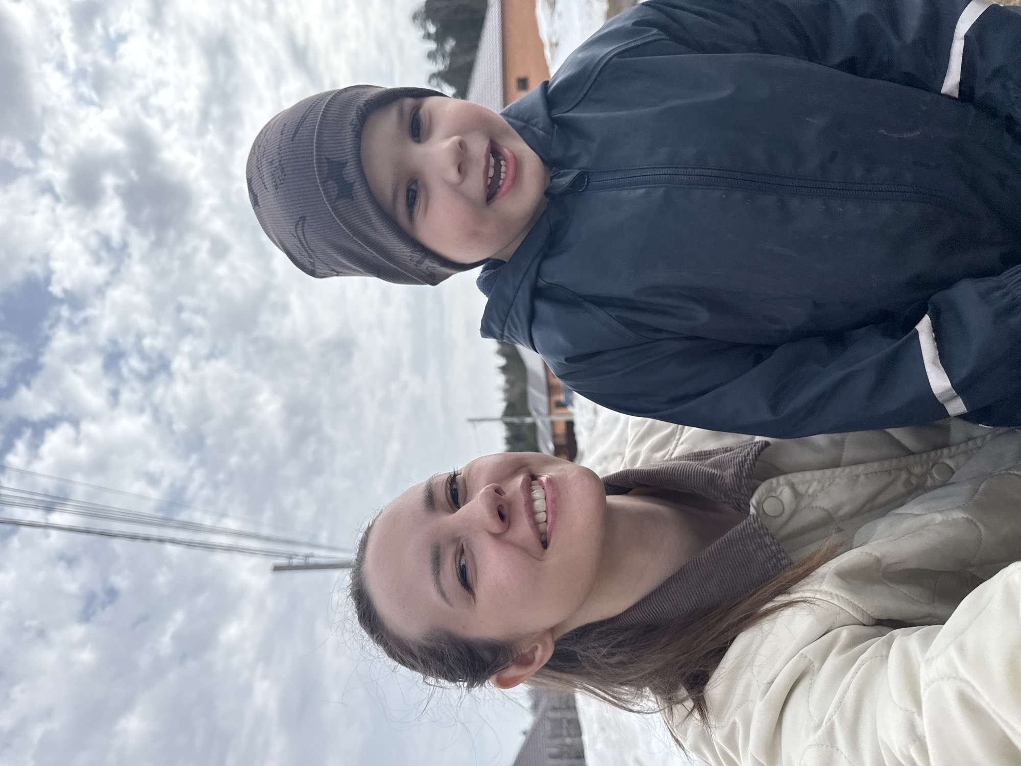 A woman and a young child are posing for a selfie outdoors on what appears to be a cloudy day. The woman is smiling at the camera. She has brown hair and is wearing a light-colored quilted jacket with a brown corduroy collar. The child, also smiling, is wearing a dark blue jacket and a gray knit hat with a pattern on it. In the background, there is a building with a brown roof and some snow on the ground. Utility poles with wires are also visible in the sky. This image was likely taken in a residential area in Chaikovsky, Russia, although specific landmarks are not discernible.