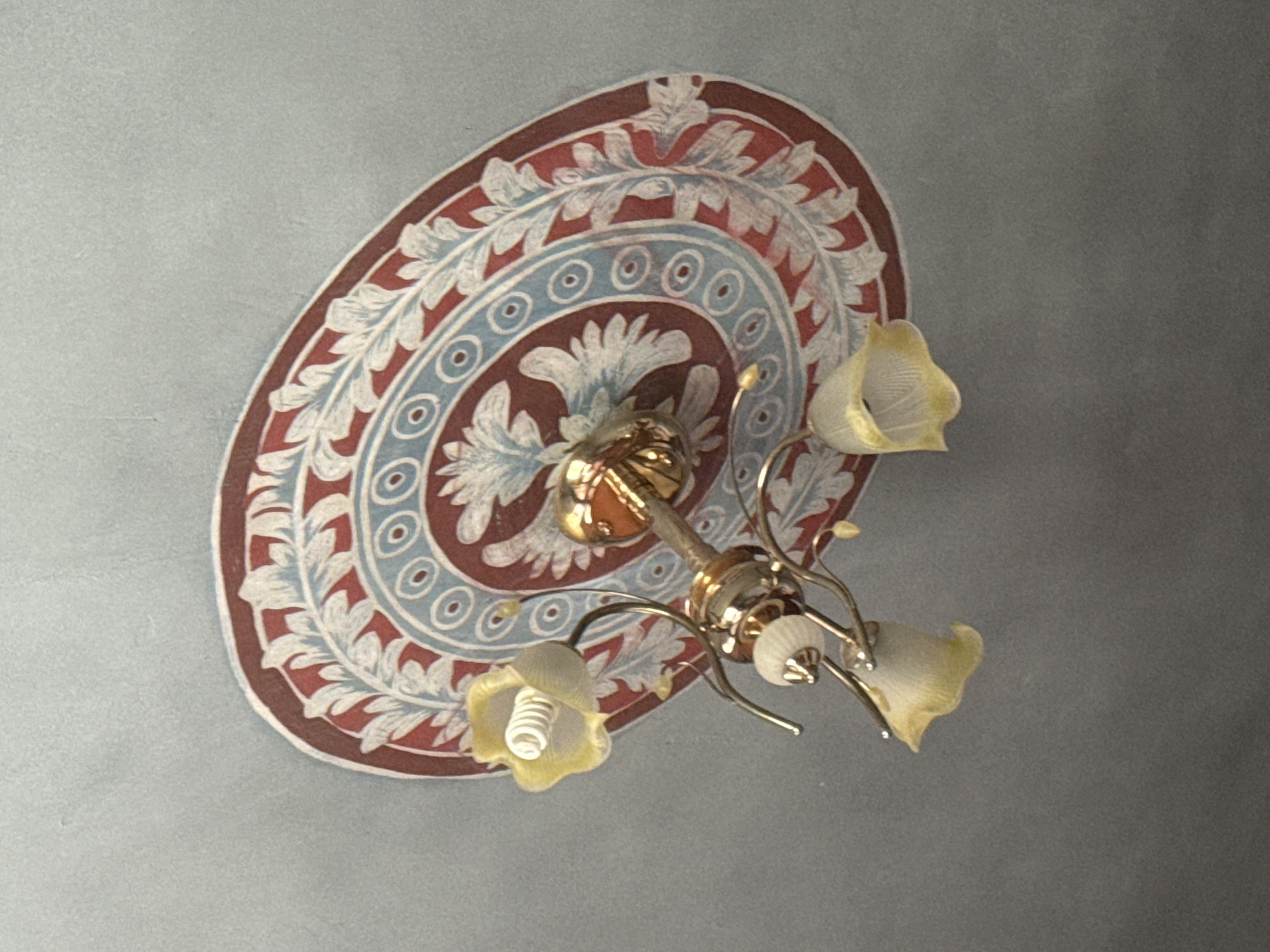 A brass chandelier with three yellow, bell-shaped glass shades hangs from a ceiling decorated with a circular fresco. The fresco features a pattern of alternating red and blue bands filled with stylized leaves and concentric circles. The chandelier's central element is a gold-colored orb, and the arms are made of thin, curved brass. Each shade contains a modern, spiral-shaped LED light bulb. The ceiling is painted a muted blue-gray. The image was taken in Kosh, Armenia, an area known for its viticulture and historical churches, though no specific landmarks are visible in this shot of a domestic interior.