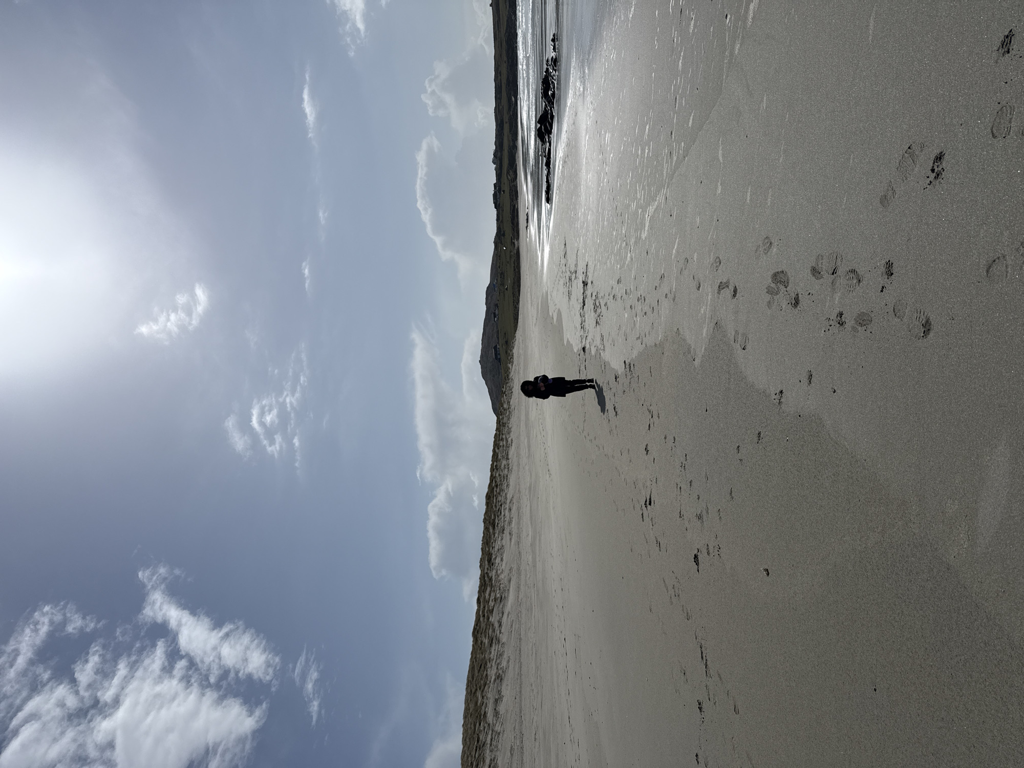 The image shows a beach scene under a partly cloudy sky. The beach has a sandy expanse with footprints visible. A person stands on the beach, their figure appearing silhouetted against the bright background. Behind them, there is a coastline with what appears to be vegetation or cliffs. The water's edge reflects light, creating a shimmering effect. The location of the scene is Unknown, Unknown.