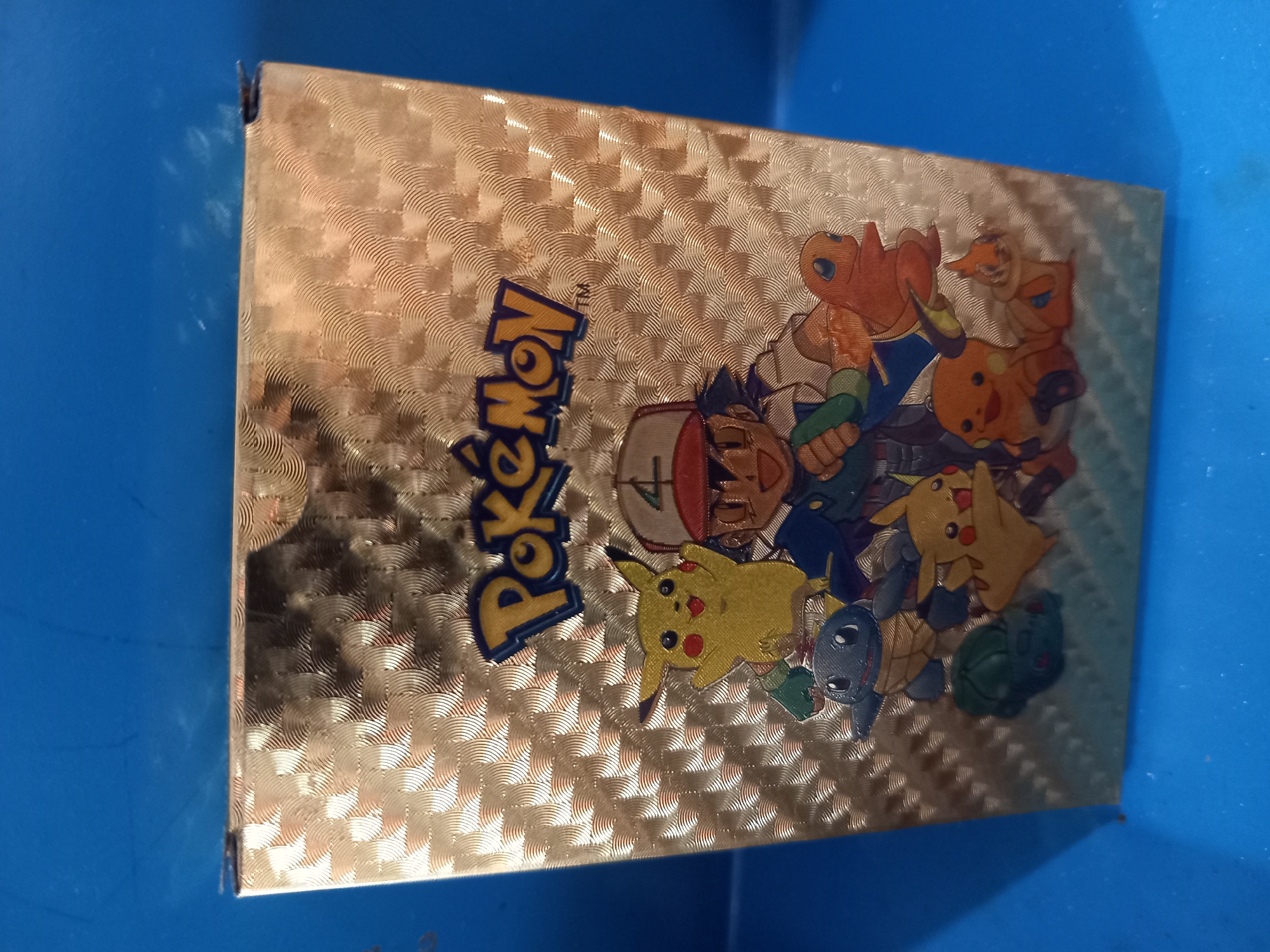 A close-up shot shows the front of a golden cigarette case adorned with a Pokemon design. The Pokemon characters, including Pikachu, Ash, Squirtle, Charmander, and Bulbasaur, are depicted in a cartoonish style. The case has a textured, patterned surface. The background is a solid blue surface. The image is taken from a slightly high angle, looking down at the case.