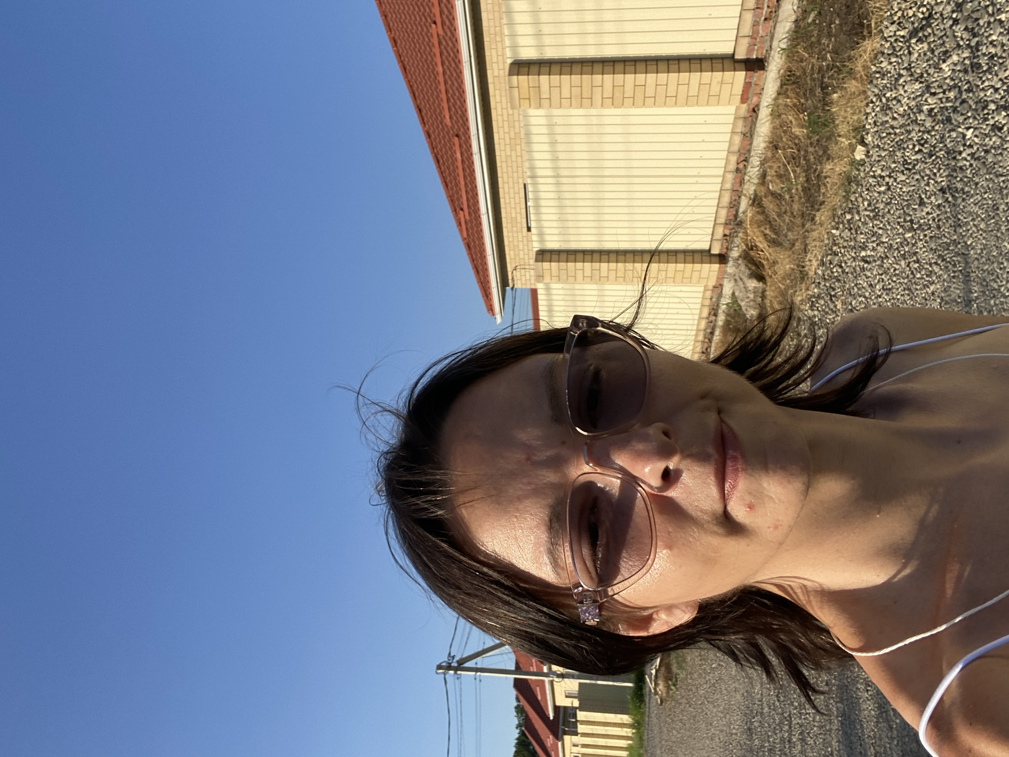 A close-up selfie of a woman with dark hair and brown eyes, wearing sunglasses, taken outdoors. She is looking towards the camera with a neutral expression. The background shows a clear blue sky, a beige fence with a red-tiled roof, and a gravel path. To the left, there are houses and a utility pole with wires. The lighting suggests it is daytime. The location is identified as Yuzhny, Russia.