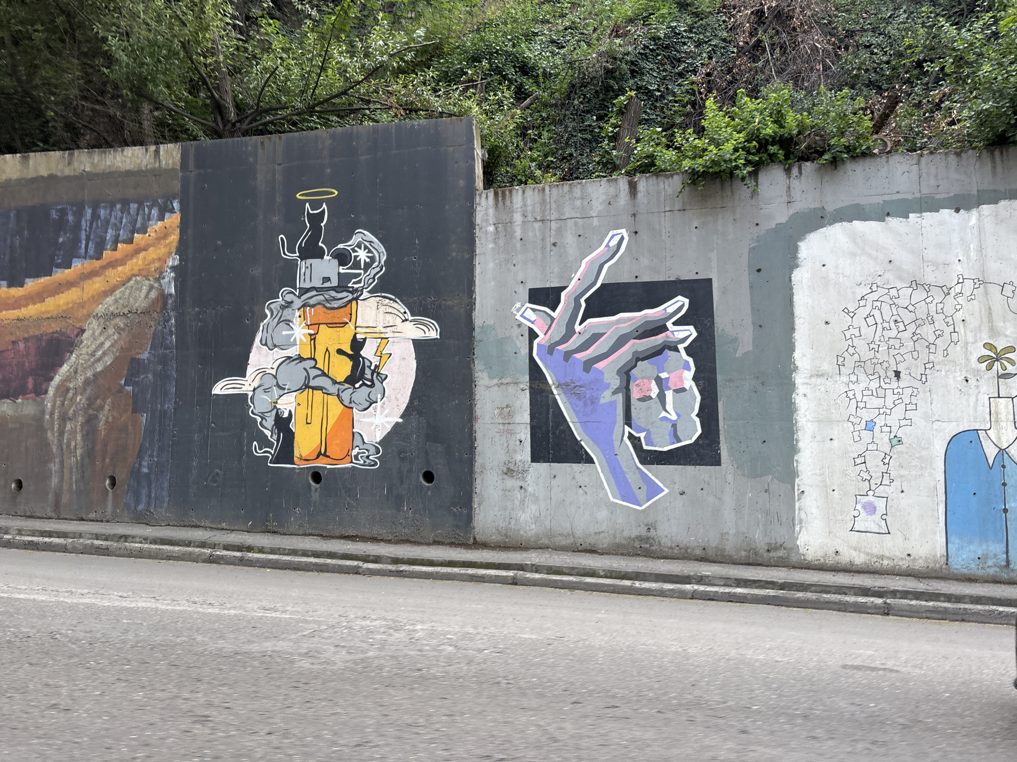 The image shows a section of a concrete retaining wall with various graffiti murals, located alongside a paved road in Tbilisi, Georgia. The murals are diverse in style and subject matter. On the left, a mural depicts what appears to be a stylized, abstract representation of a staircase or ramp with orange and brown tones. To its right, a black mural features a whimsical drawing of a black cat with a halo, sitting atop a cartoonish, yellow structure with smoke or clouds emanating from it, along with lightning bolts and gears. The center mural is a graphic depiction of a hand with extended fingers, rendered in shades of purple, pink, and grey against a black background. Further to the right, on a lighter grey section of the wall, is a more abstract mural with a network of interconnected squares and lines, with a few colored squares, and a stylized plant with a green head. The far right of the visible wall features a portion of a figure wearing a blue jacket and a white head. The retaining wall itself appears weathered, with some staining and discoloration. Vegetation, including trees and ivy, grows above the wall on a sloped embankment. The road in the foreground is asphalt, with a curb and gutter. The lighting suggests it is daytime.