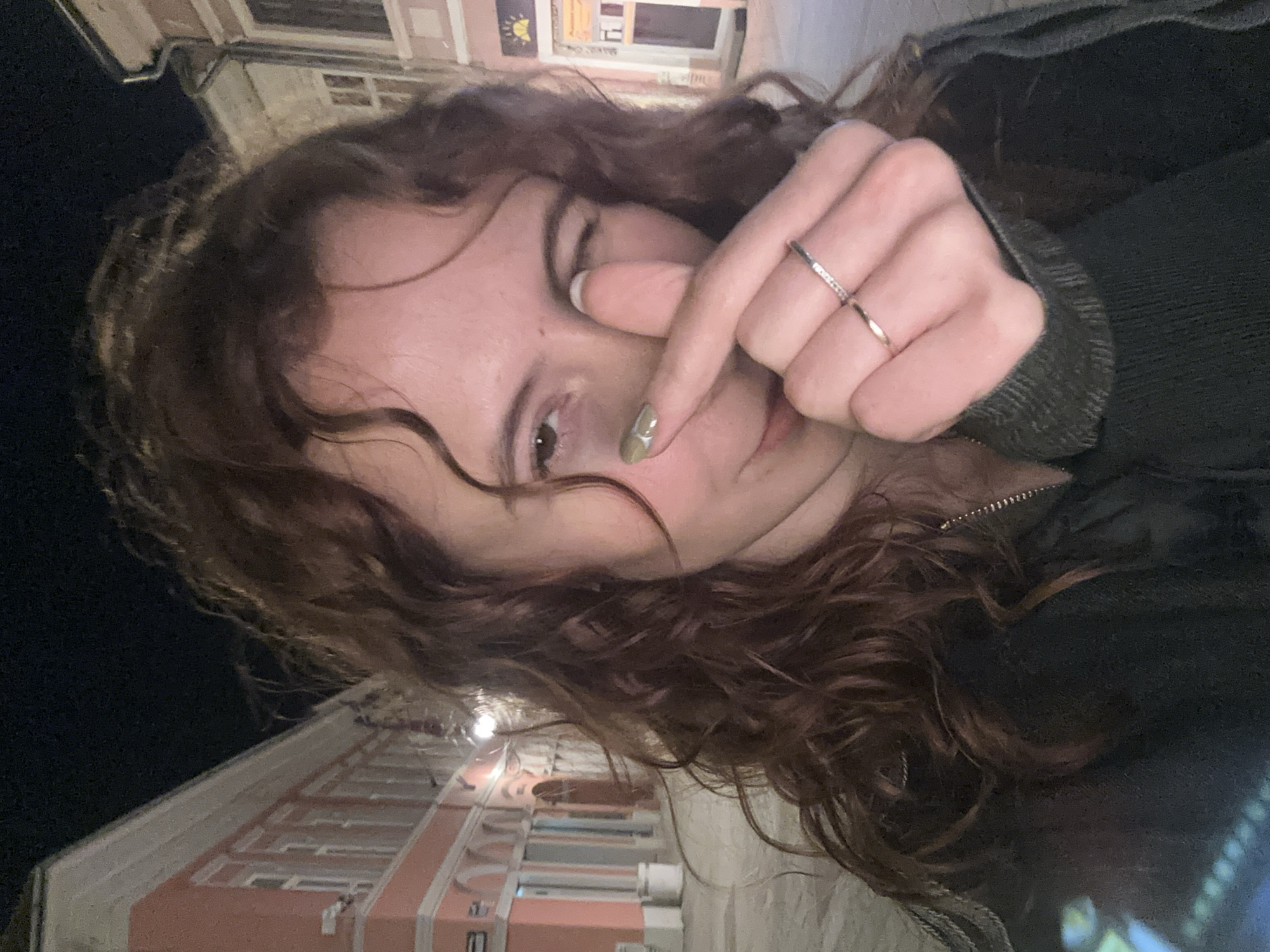 A woman with curly brown hair is taking a selfie outdoors at night. She is wearing a dark-colored jacket and two rings on her right hand. Her left index finger is pointing towards her eye, and her fingernail is painted light green. In the background, there is a building with pink and white architectural details, indicative of a European-style facade. Based on the architectural style, the location is likely in the city center of Novi Sad, Serbia.