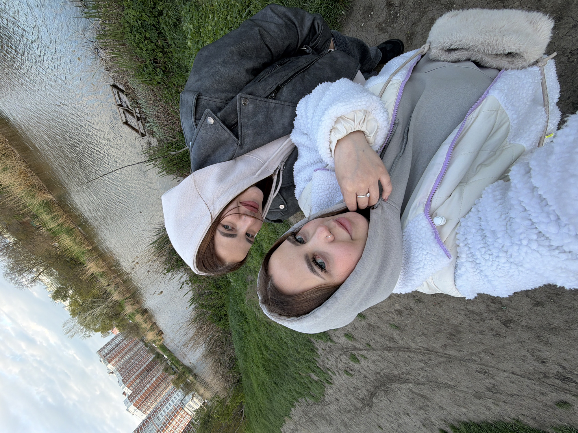 The provided image, which is oriented with the horizon running vertically, features two young women captured in a close-up outdoor selfie, looking directly at the camera. They both wear hooded tops with their hoods up and display neutral to gently smiling expressions.
The woman on the left is dressed in a light beige hooded garment underneath a dark grey or black textured jacket. The woman on the right wears a light grey hooded top, overlaid with a fluffy white jacket that has visible lavender zippers and interior lining details. She has a small hoop piercing in her right nostril and a silver-colored ring on the ring finger of her left hand. Her eyes are blue/green.
They are positioned on a strip of dirt and patchy green grass. Alongside them is a body of water, likely a river or canal, showing ripples on its surface. A small wooden structure or piece of debris is partially visible in the water. On the opposite bank, there are dry reeds and trees, many of which have bare branches, suggesting late winter or early spring.
In the background, across the water, stand several tall residential buildings characterized by red/orange and white facades, set against a partly cloudy sky. The scene depicts an urban or suburban waterfront area, aligning with the context of Krasnodar, Russia, photographed during daytime under soft, diffused lighting conditions. There is no visible text in the image.