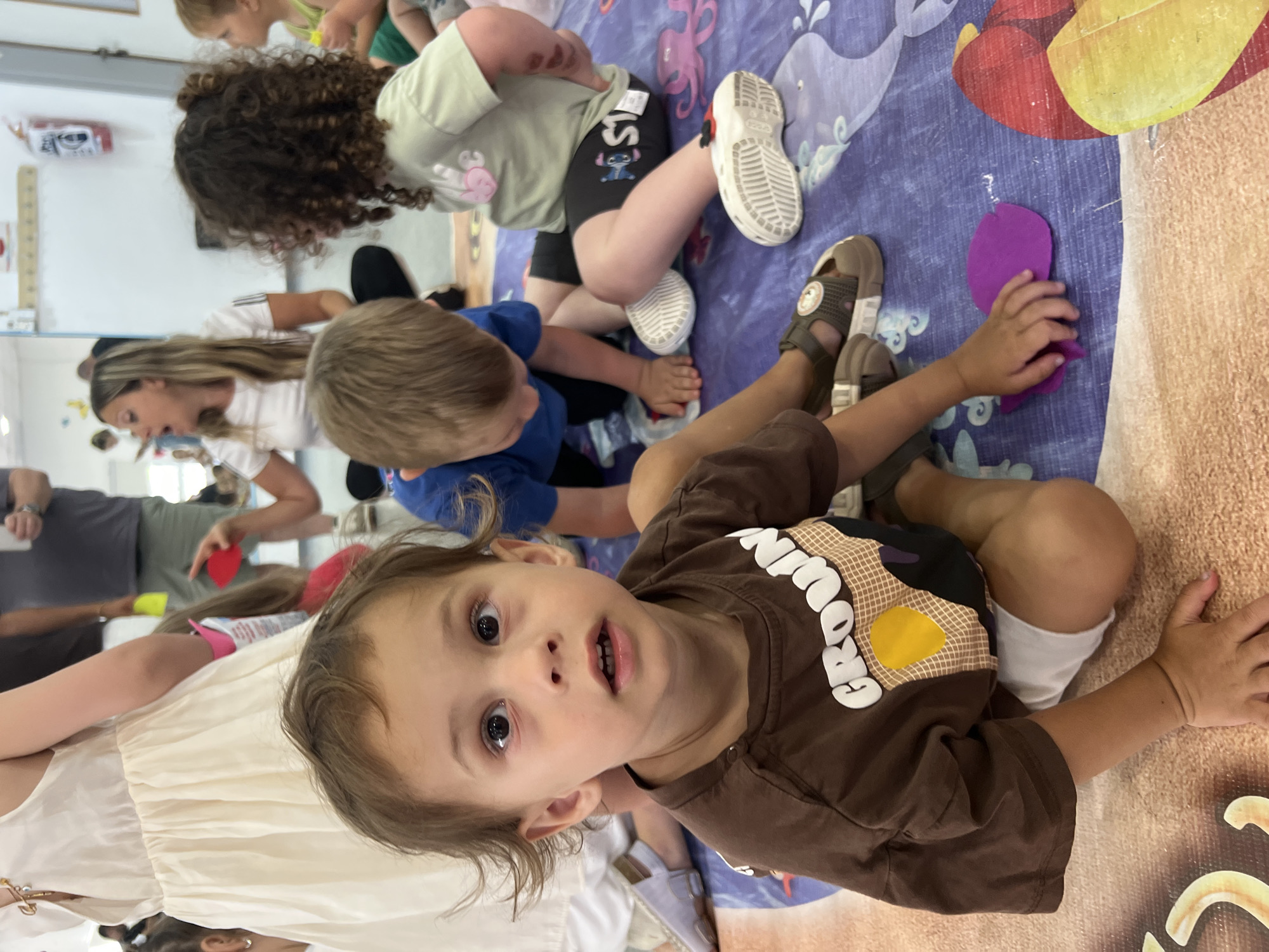 The image shows a group of young children and adults in a brightly lit, indoor setting. The floor is covered with a large, colorful mat featuring sea creatures and waves, suggesting a play area, possibly a daycare or a children's activity center. The children appear to be engaged in some sort of activity, possibly craft or play, as some are sitting and interacting with materials on the mat.
In the foreground, a young child with large, dark eyes and brown hair is looking directly at the camera, with an open-mouthed expression that could be surprise or excitement. They are wearing a brown t-shirt with white text that reads "BROWNIE" and a yellow shape on the chest. To their left, another child in a blue shirt is partially visible, and further to the left, an adult with long blonde hair is also present.
Other children are seen sitting on the mat. One child has curly dark hair and is wearing a light green t-shirt and dark shorts with a Stitch character printed on them, along with white Crocs. Another child's legs are visible, wearing sandals. A purple cutout, possibly made of felt or paper, is being touched by a child's hand on the mat.
The overall atmosphere seems lively and playful. The lighting suggests it is daytime. The presence of mats, toys, and multiple children and adults indicates a communal activity for young children. There is no indication of the weather or time of day beyond it being indoors during daylight. The location context is Kiryat Bialik, Israel.