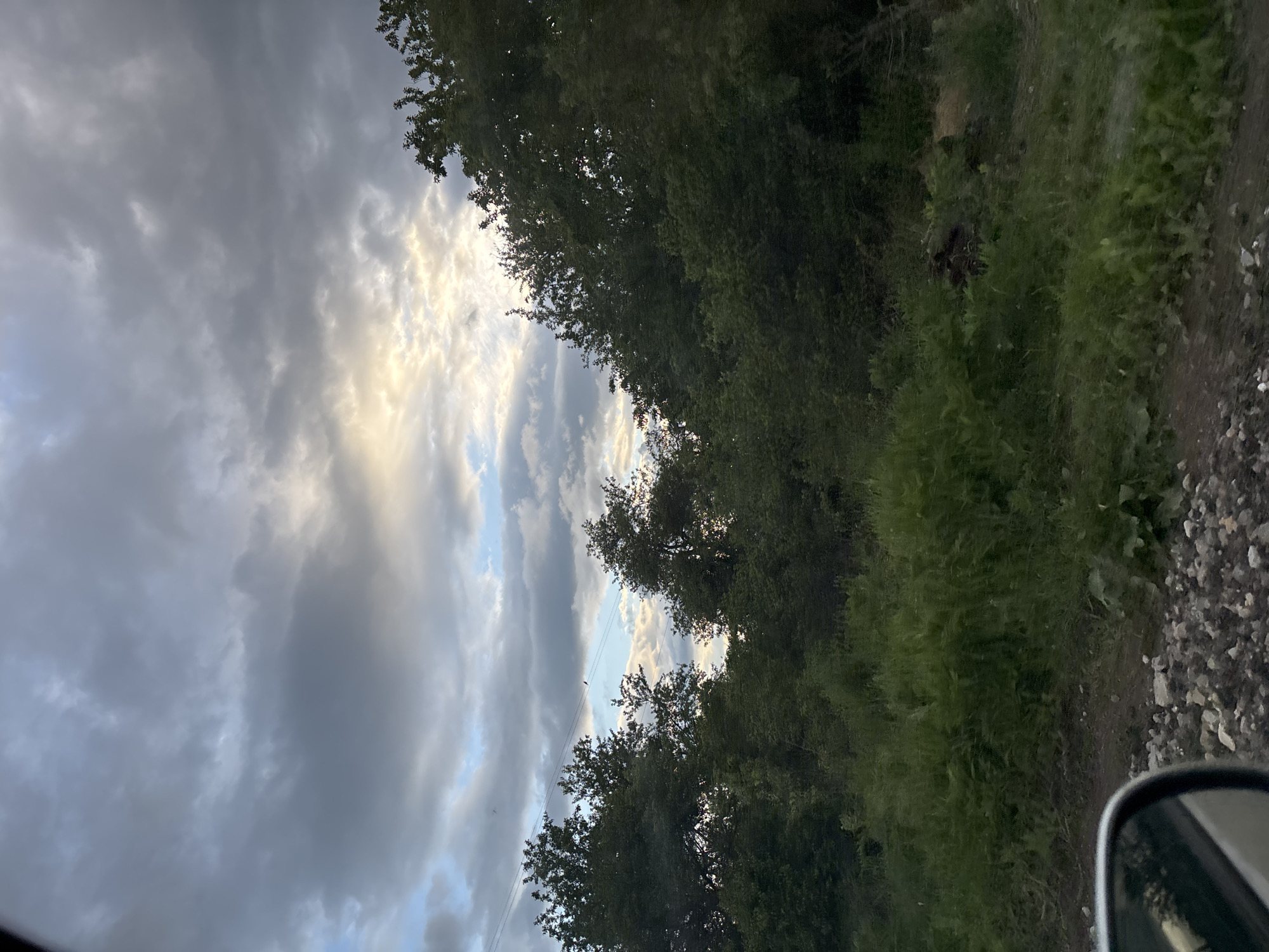 The image captures a view from a vehicle in what appears to be a rural location near Baylovo, Bulgaria. The sky is heavily overcast with a mix of dark and light clouds, with a patch of light filtering through. Trees line the right side of the frame, with dense green foliage. A dirt or gravel road runs alongside the trees, transitioning to a rocky area. The side mirror of the vehicle is visible in the bottom right corner.
