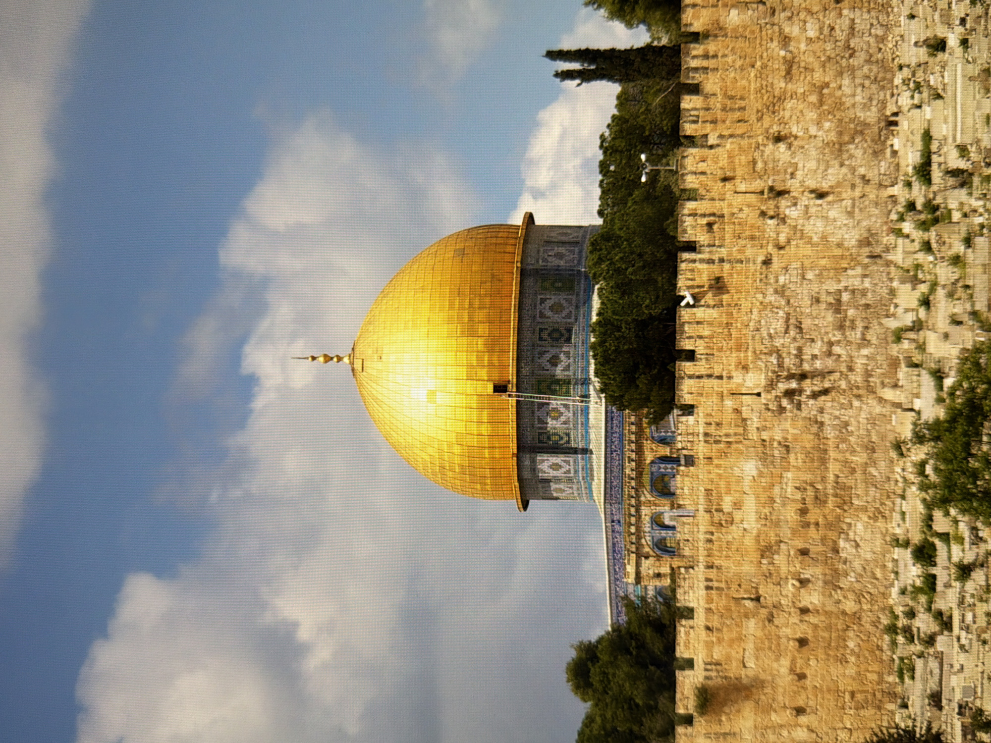 The image depicts the Dome of the Rock, a sacred Islamic shrine located in Jerusalem. The golden dome, a prominent feature, is situated atop a cylindrical drum adorned with intricate mosaic tilework. Below the dome, a stone structure with arched windows is visible. The shrine is surrounded by lush green trees and stands behind a massive, ancient stone wall. The wall, constructed from large, rough-hewn blocks, is weathered and shows signs of age. The foreground of the image reveals a terraced slope covered with smaller stones and sparse vegetation, possibly a graveyard or an ancient archaeological site. The sky above is a vibrant blue, with scattered white clouds. The overall scene is one of historical and religious significance, capturing the architectural grandeur of the Dome of the Rock and its surrounding ancient landscape. The location is Jerusalem, not Kyiv, Ukraine.