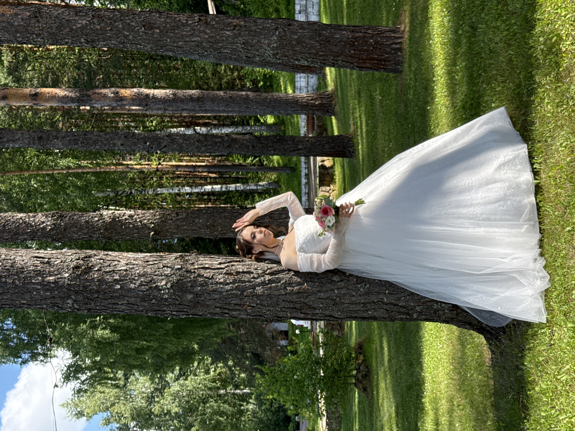 A bride stands in a sun-dappled forest clearing near Votkinsk, Russia. She is leaning against a large tree, her white off-the-shoulder wedding dress flowing around her. Her right hand is raised to shield her eyes from the sun, and she holds a bouquet of pink and white flowers in her left. The background features more trees, with a glimpse of a building and a stone wall in the distance. The ground is covered in green grass, with shadows cast by the trees. The sky above is partly cloudy.
