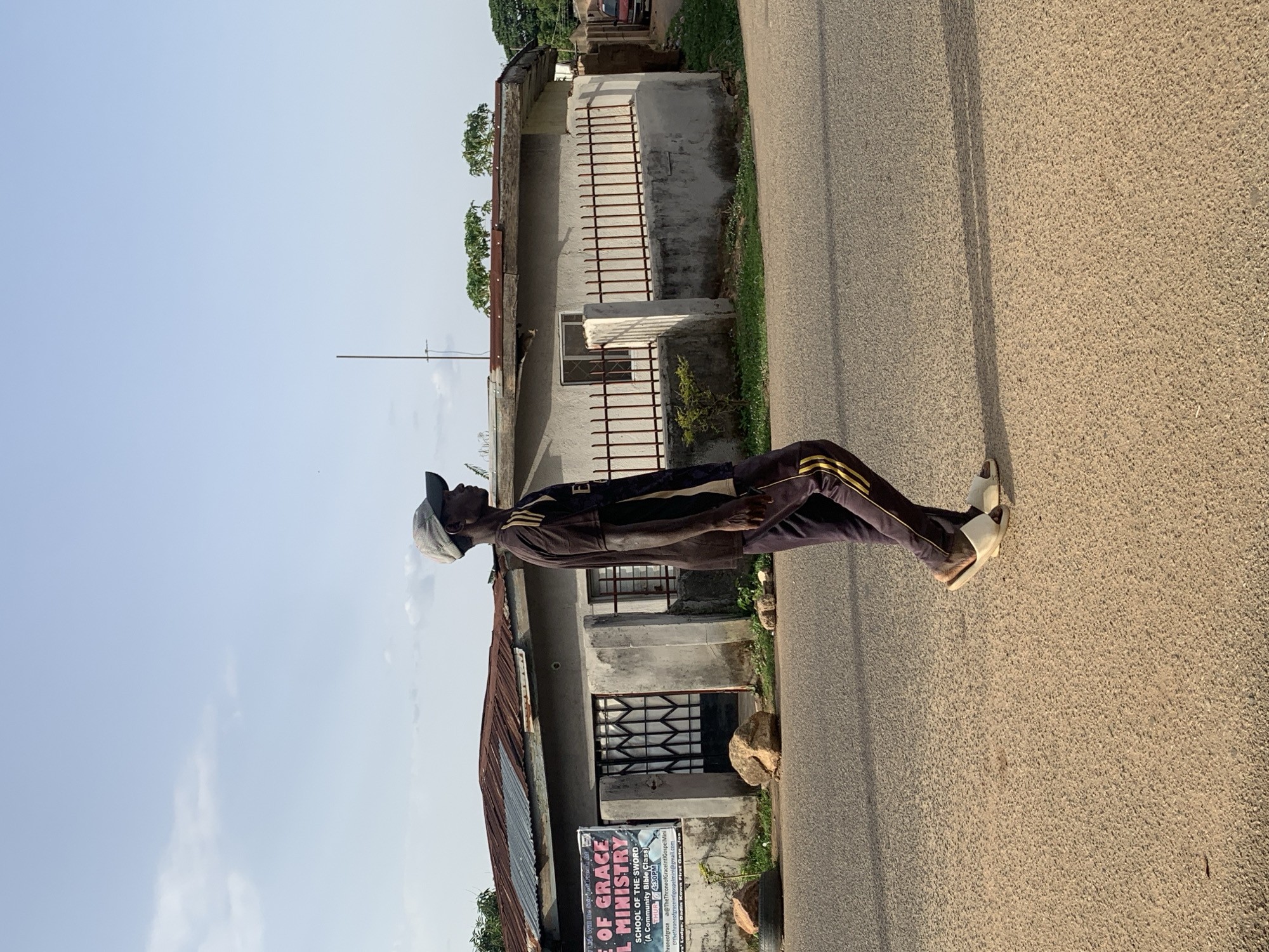 A medium shot, oriented vertically, depicts a man standing on a paved road, with a building in the background.
The man is facing the left of the frame, wearing a cap, a brown shirt, dark-colored pants with gold stripes, and sandals.
Behind him, a weathered concrete building with barred windows is visible. A sign for the "Throne of Grace Eternal Ministry" hangs on the building's exterior. The sign is written in English.
The scene is lit by natural light.