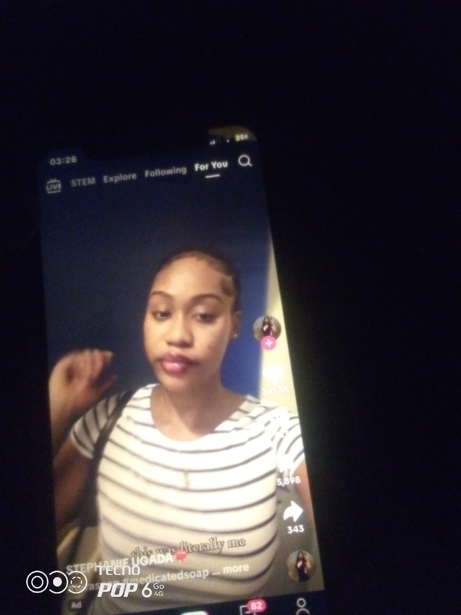 The image is a screenshot of a TikTok video on a smartphone. The video features a young woman with dark skin and braided hair, wearing a striped shirt. She is looking at the camera and appears to be speaking. Text overlays on the screen indicate the username "STEPHANIE UGADA" and a comment that reads "this was literally me". There are also icons for likes, shares, and comments, as well as a profile picture of another user. The background is dark and out of focus. The phone itself appears to be a Tecno Pop 6 4G. The time on the phone's screen is 03:26. The city of Howayi, Nigeria is not mentioned in the image and cannot be determined from the visual information.
