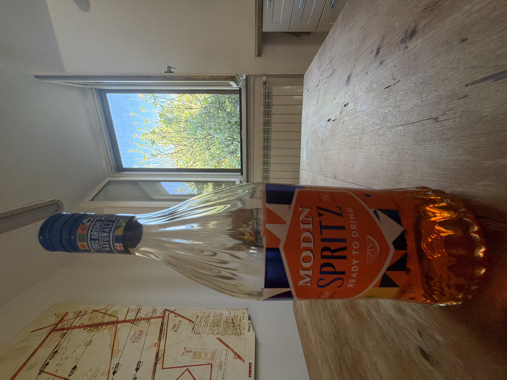 The image shows a bottle of Modin Spritz lying on a wooden table. A map of Brazil, specifically Sao Paulo, is partially visible under the bottle. A window in the background is open, revealing trees and the sky outside. A radiator is visible near the window. The city of Milan, Italy, is not apparent in this image, as the map suggests a Brazilian location.