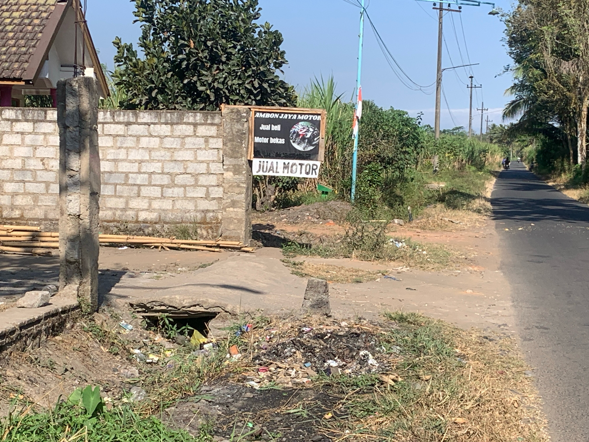 This is a full, eye-level shot of a rural roadside scene in Sukonolo, Indonesia. The foreground features a ditch with green grass and a pile of assorted trash, including burnt material, plastic, and paper. A concrete structure, possibly a broken drain cover, is situated in the ditch. To the left, a concrete wall made of light-colored bricks stretches across the frame. Behind the wall, a portion of a building with a brown tiled roof is visible, along with some bamboo poles stacked near the wall.
Adjacent to the wall, a wooden sign with black text reads "AMBON JAYA MOTOR" at the top, followed by "Jual beli Motor bekas" and an image of motorcycles. Below this, a larger white sign displays the text "JUAL MOTOR".
To the right of the sign, a light blue utility pole with electrical wires extends upwards, with a small Indonesian flag attached to it. Beyond the sign and pole, dense green vegetation and trees line the roadside. A paved road runs from the middle ground into the distance, where two individuals on motorcycles are visible. The sky is clear and blue. The overall impression is that of a simple, open-air business operating on the edge of a rural community.