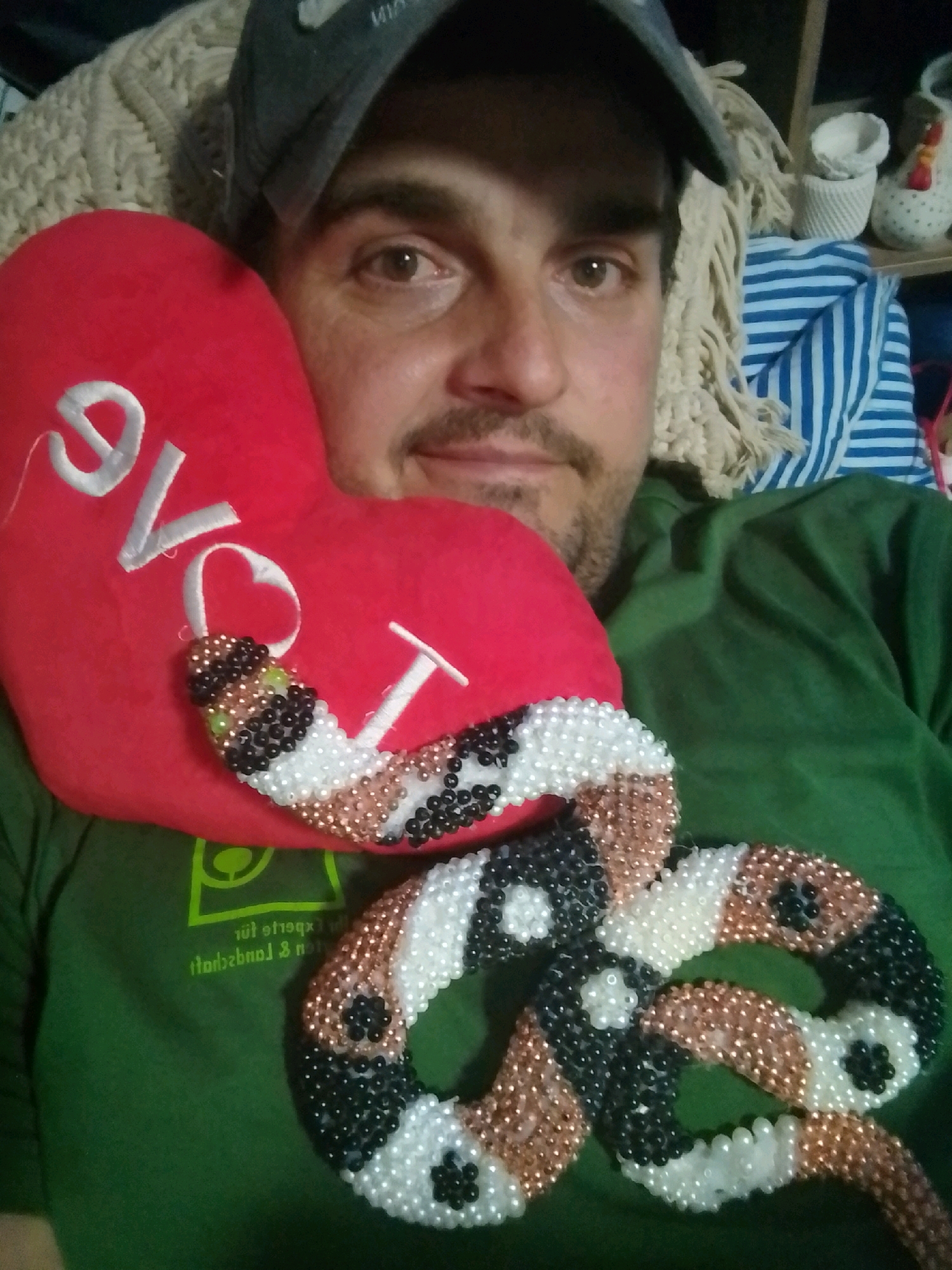 A man, wearing a green t-shirt and a baseball cap, is lying down with a red heart-shaped pillow and a beaded snake. The pillow has "Love" written in white cursive letters. The beaded snake is wrapped around the pillow and extends down the man's chest. The background shows a knitted blanket and some decorative items on a shelf. There is no discernible information about the location in Wedel, Germany within the image.