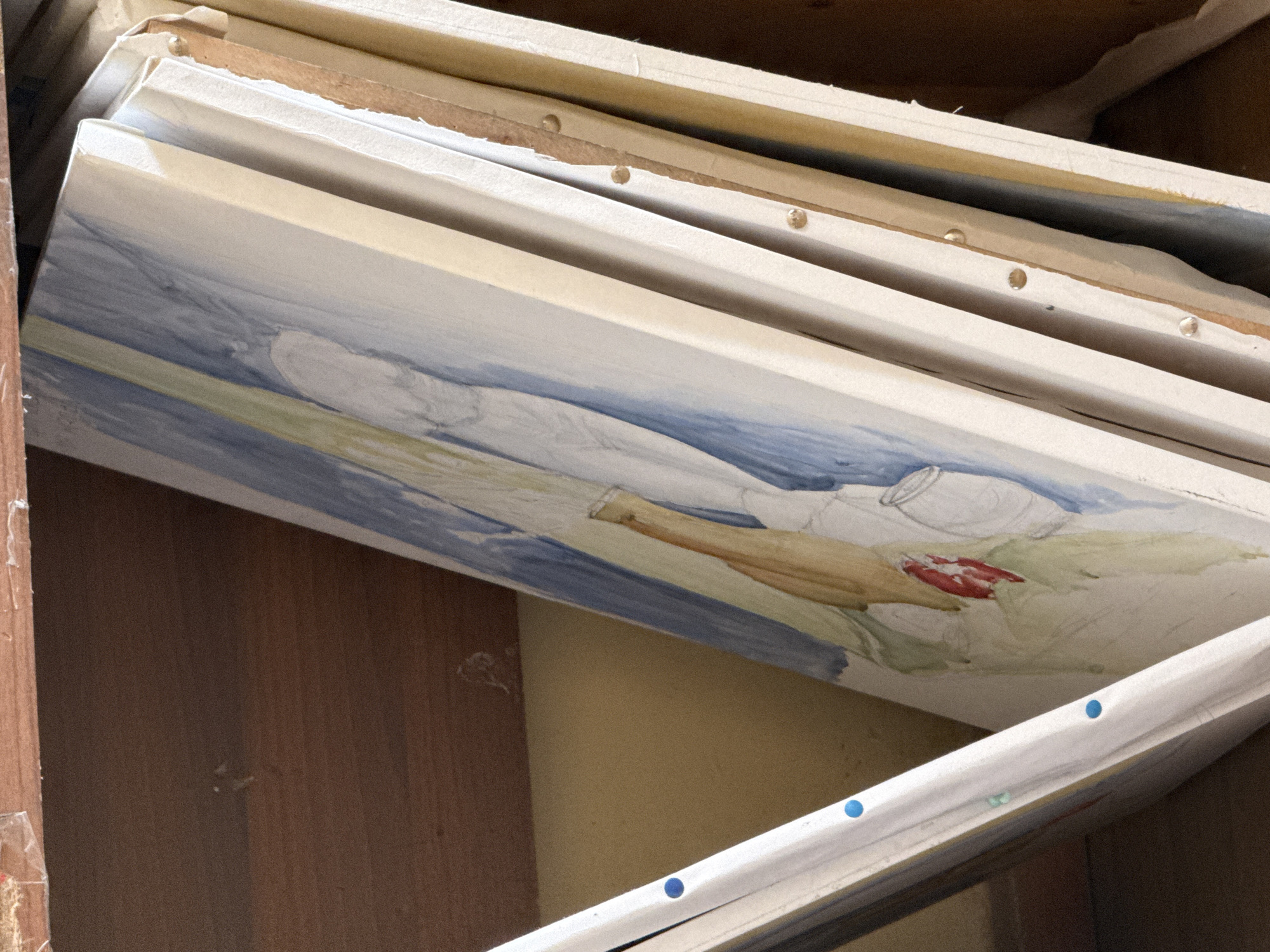 The image shows a stack of partially painted canvases leaning against the interior walls of a storage space. The artwork visible depicts a still life composition with a white cloth, vase, and possibly some fruit. The paint is applied in broad strokes of blue, green, brown, and red. The canvases are framed and stacked at an angle, with bare wood and staples visible along the edges. The storage area appears cluttered, with wood panels and other unseen items surrounding the canvases. There is no information about Yerevan, Armenia, or any people.