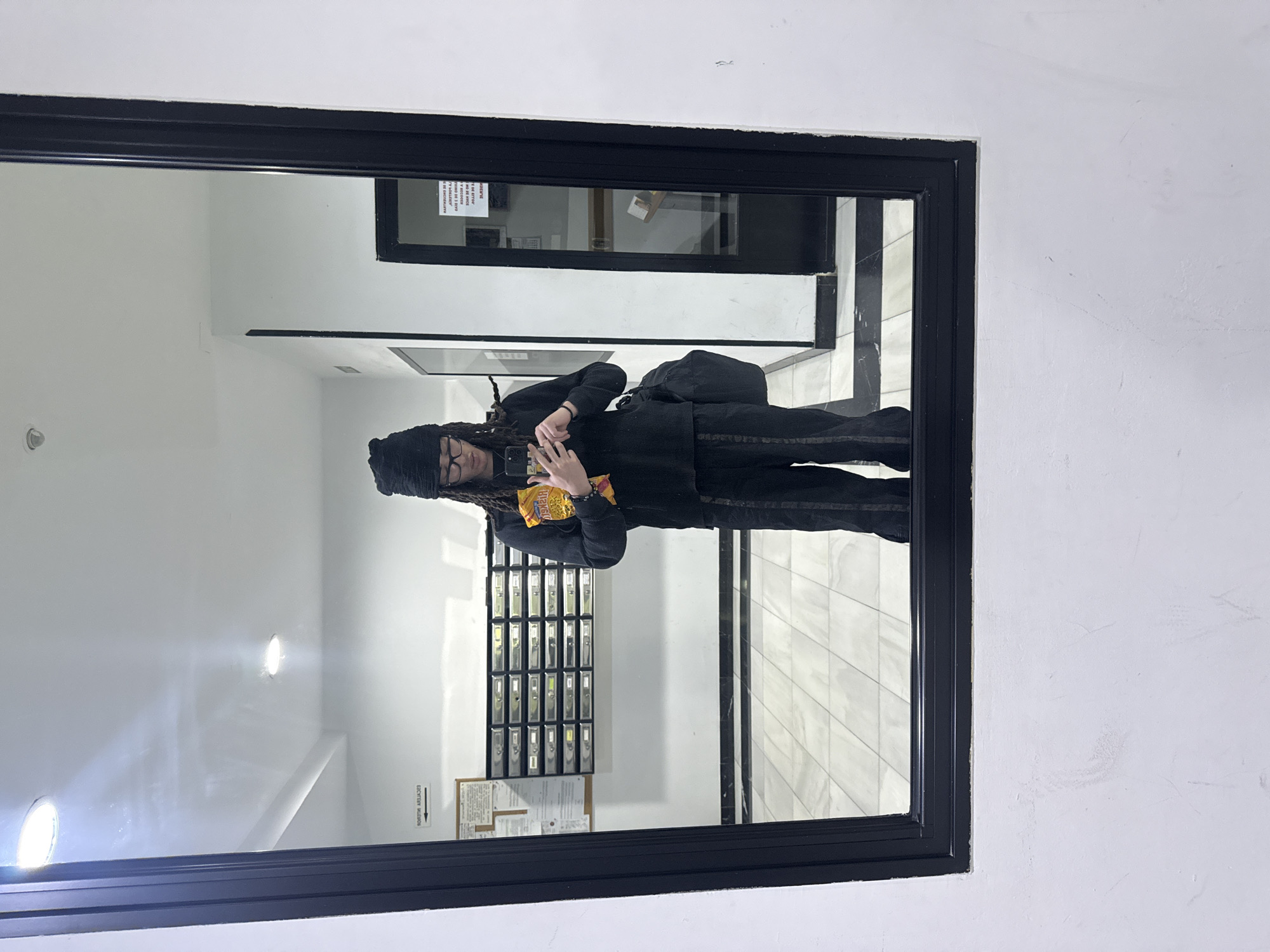 A mirror selfie captures a person standing in what appears to be an indoor hallway or lobby, likely in an apartment building in Madrid, Spain. The individual, dressed in a dark, long-sleeved top, dark pants, and wearing glasses with a dark beanie, holds a smartphone to take the photo. They have prominent dreadlocks cascading from under their beanie. In their other hand, they hold a yellow bag of snacks, possibly chips, with the word "Habanero" visible on the packaging. A dark shoulder bag is worn over their left shoulder.
The large rectangular mirror, encased in a thick black frame, reflects a bright, clean-lined interior. Behind the person in the reflection, there is a wall-mounted mailbox system featuring multiple individual metal mailboxes. Below these mailboxes, an informational bulletin board with text is partially visible, reading "BOLETIN DE AVISOS" (Notice Board) in Spanish, along with other building-related information. Another framed document with Spanish text is visible higher up in the reflection.
The walls of the hallway are white, and the floor appears to be a light-colored, possibly marble-patterned, tiled surface. Several circular recessed ceiling lights brightly illuminate the space. The overall scene suggests a functional and modern entryway, common in residential or commercial buildings. The person's expression is neutral, focused on capturing the image. The artificial lighting inside means no specific time of day can be determined.