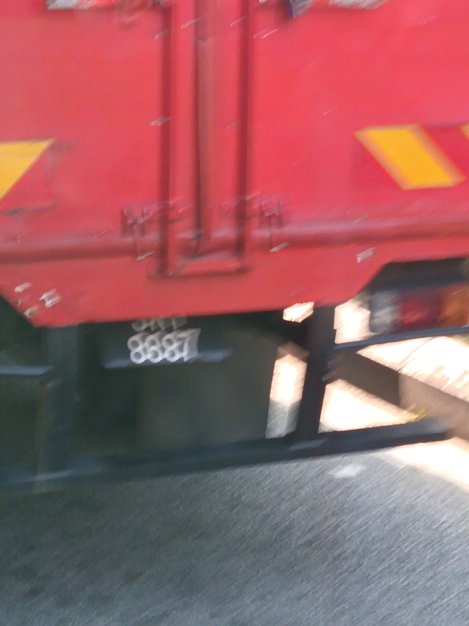 The image shows a close-up, blurry view of a red train car. On the lower part of the train car, a black rectangular plate is visible with the number "8887" stenciled in white. The train car has yellow reflective triangles on its side. The ground in front of the train is a textured, grey surface, possibly asphalt. The overall scene is out of focus, suggesting movement or a low-quality photograph. No specific location in Kuala Lumpur, Malaysia can be determined from the image.