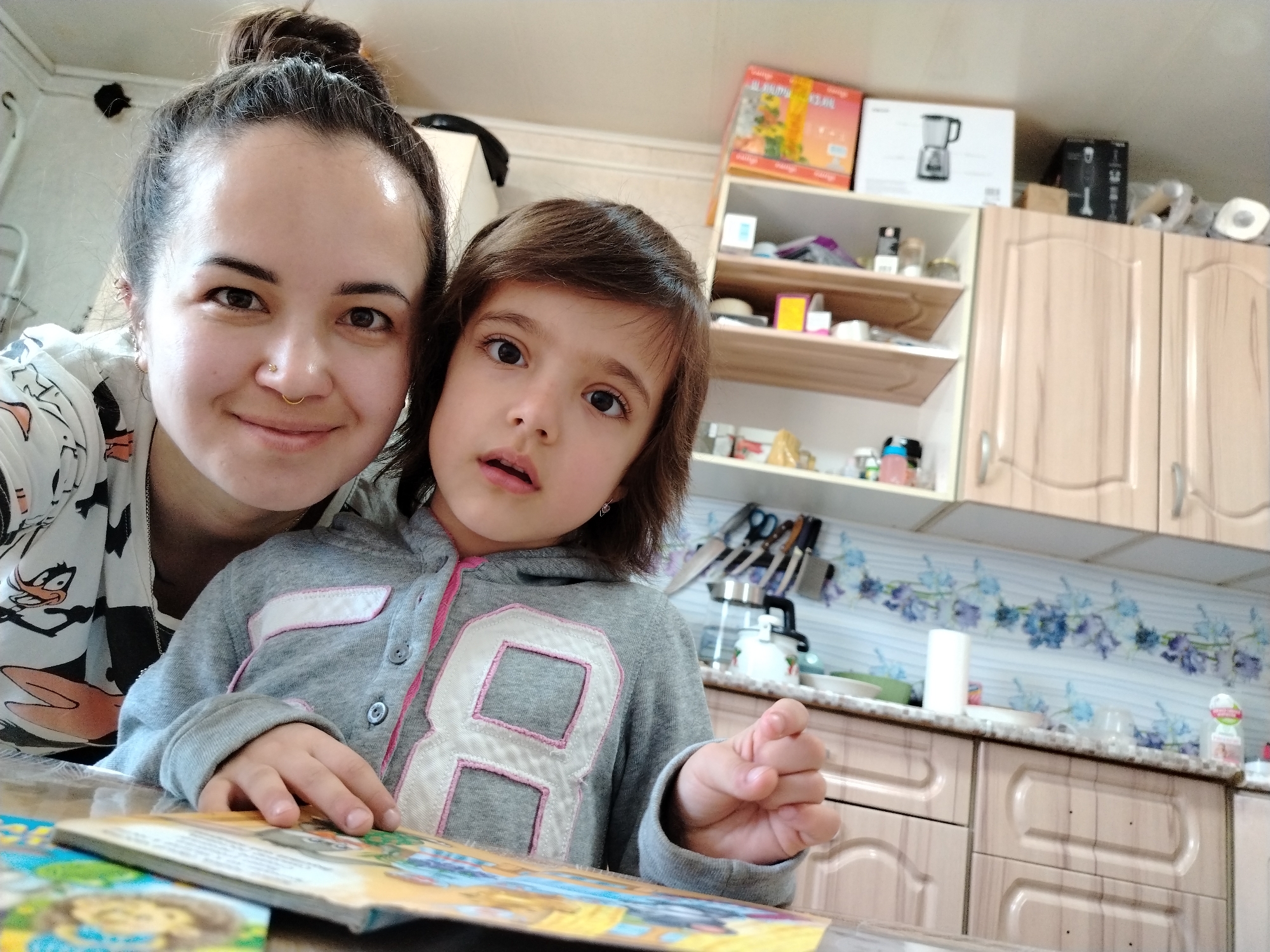 A woman with her hair in a bun, wearing a patterned top, stands beside a young girl in a gray hooded sweatshirt with a large white "8" on the front. They are in a kitchen in Гожан, Russia, with light brown cabinets and shelves. The woman is looking at the camera and smiling, while the girl is looking to the side with a slight expression. A book lies open on the table in front of them. Kitchen utensils, appliances, and boxes are visible on the shelves and countertops.