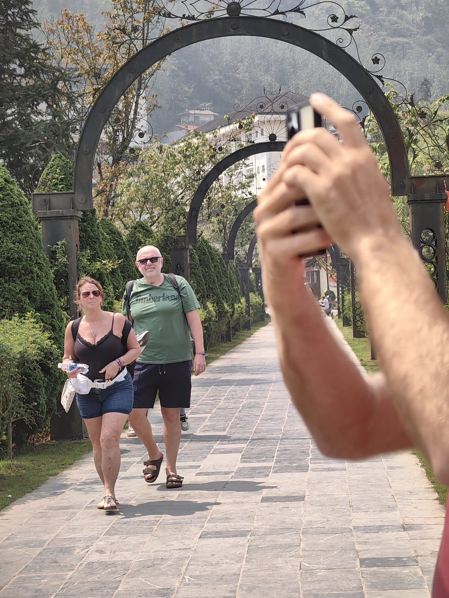 The image captures a paved pathway in Sa Pa, Vietnam, extending into the background beneath a series of dark, ornate metal arches. In the immediate foreground, the arms and hands of a person hold a mobile phone horizontally, pointing it towards the midground subjects.
Two individuals are walking on the pathway towards the viewer. The female on the left wears a black tank top, denim shorts, and light-colored flat sandals. She has sunglasses on, a dark backpack, and a white fanny pack, and carries a clear plastic water bottle along with a white patterned item. The male on the right wears a green "Timberland" t-shirt, dark shorts, and dark brown two-strap sandals. He also wears sunglasses and a dark backpack. Both individuals are walking side-by-side.
The pathway is composed of rectangular grey paving stones. On either side, manicured green bushes and grass strips flank the path, leading up to the decorative metal arches. Beyond the arches, a landscape of green trees and hazy hills is visible, interspersed with buildings, one of which exhibits a tiered roof design typical of traditional Asian architecture. The scene is illuminated by direct sunlight, casting shadows on the ground.