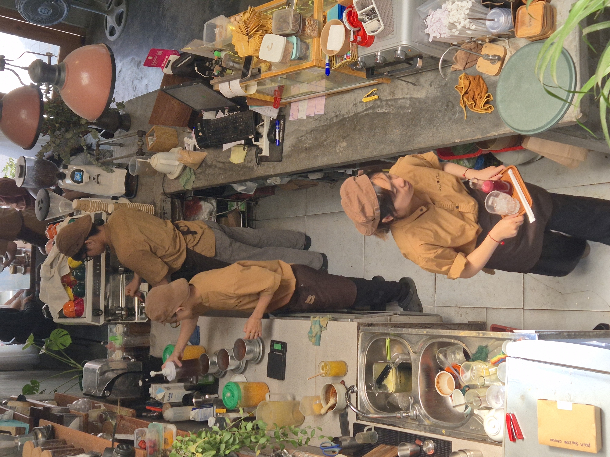 The image captures a high-angle, well-lit view of a busy cafe counter in Da Nang, Vietnam. Two baristas, both wearing brown aprons and caps over brown shirts, are actively working behind the counter. The barista on the left is pouring liquid from a tall bottle into a metal container, while the barista on the right is holding a tray with a glass of pink liquid and a straw.
The counter is cluttered with various cafe equipment and supplies. To the left, a stainless steel sink is filled with cups and cleaning supplies. To the right, a coffee machine, grinder, and various bottles and containers are visible. Customers are partially visible in the background, seated at tables. The overall environment appears to be a typical cafe setting with a focus on beverage preparation.