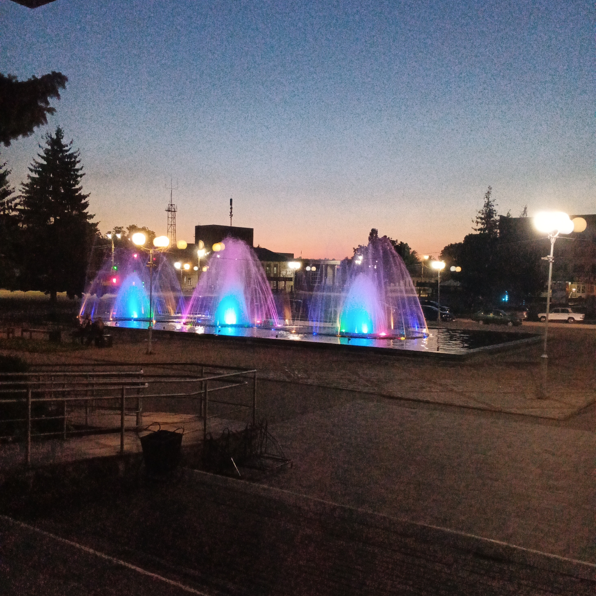 The image depicts a public square at dusk in Smila, Ukraine, featuring a multi-jet fountain illuminated with vibrant blue and pink lights. The fountain is the central focus, with water streams arching upwards and outwards, creating a dynamic visual display against the twilight sky. Several streetlights with multiple globes are visible, adding to the ambiance and illuminating the surrounding area. In the background, buildings and trees form a silhouette against the fading light. There appear to be two individuals sitting on a bench near the fountain. The foreground shows a paved walkway and a grassy area with some park amenities, including a dark-colored basket and some fencing. Several cars are parked in the distance, and a tall communication tower is visible on the skyline. The overall scene suggests a well-maintained public space designed for evening recreation.